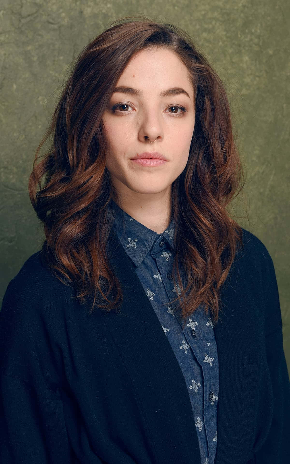 Olivia Thirlby 桌布 壁紙 — 一位年輕女性，波浪棕色頭髮和鼻環，在質感橄欖綠背景前直視鏡頭。