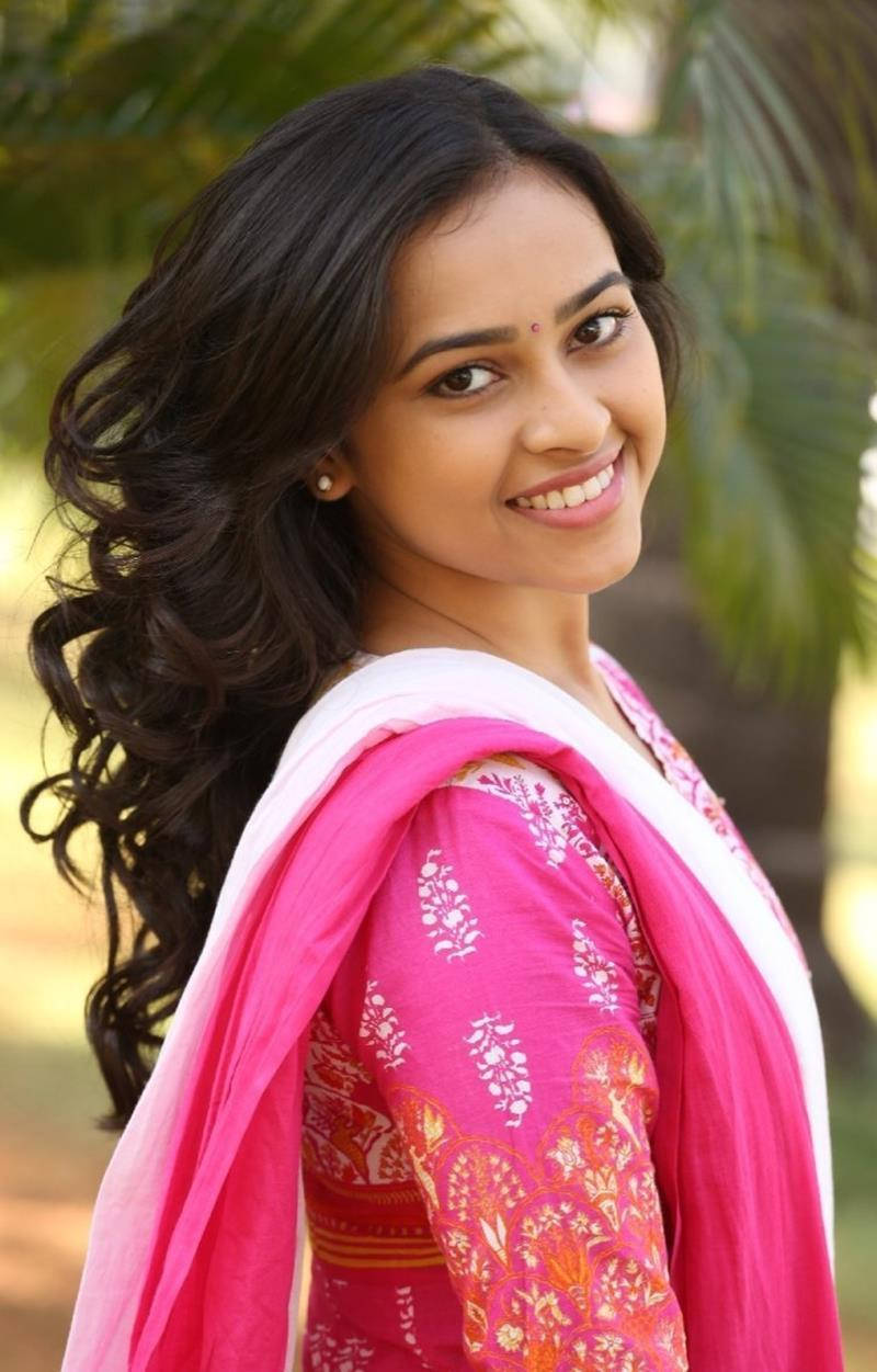 Tamil Actors HD 桌布 壁紙 — 一位年輕女性，深色波浪長髮，穿著鮮豔的粉白圖案傳統服裝，在模糊的綠色棕櫚樹背景下溫暖微笑。