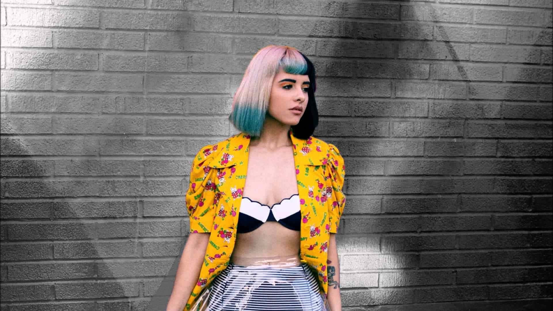 Melanie Martinez 桌布 壁紙 — 一位年輕女性，粉紅色和藍色的頭髮，穿著大膽的黃色花卉外套，背景是灰色磚牆。