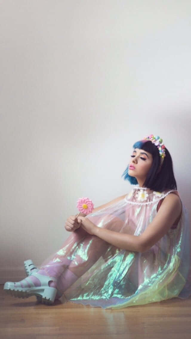 Melanie Martinez 桌布 壁紙 — 一位年輕女性，藍色頭髮，戴著花冠，穿著彩虹色連衣裙，坐在木地板上，手持粉紅色花朵，靠著素色牆壁。