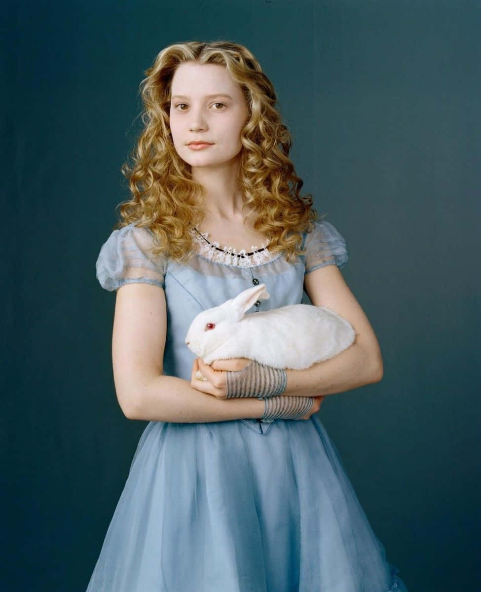 Mia Wasikowska 桌布 壁紙 — 一位年輕女性，金色捲髮，身穿淺藍色連衣裙，在深藍綠色背景前抱著一隻白兔。