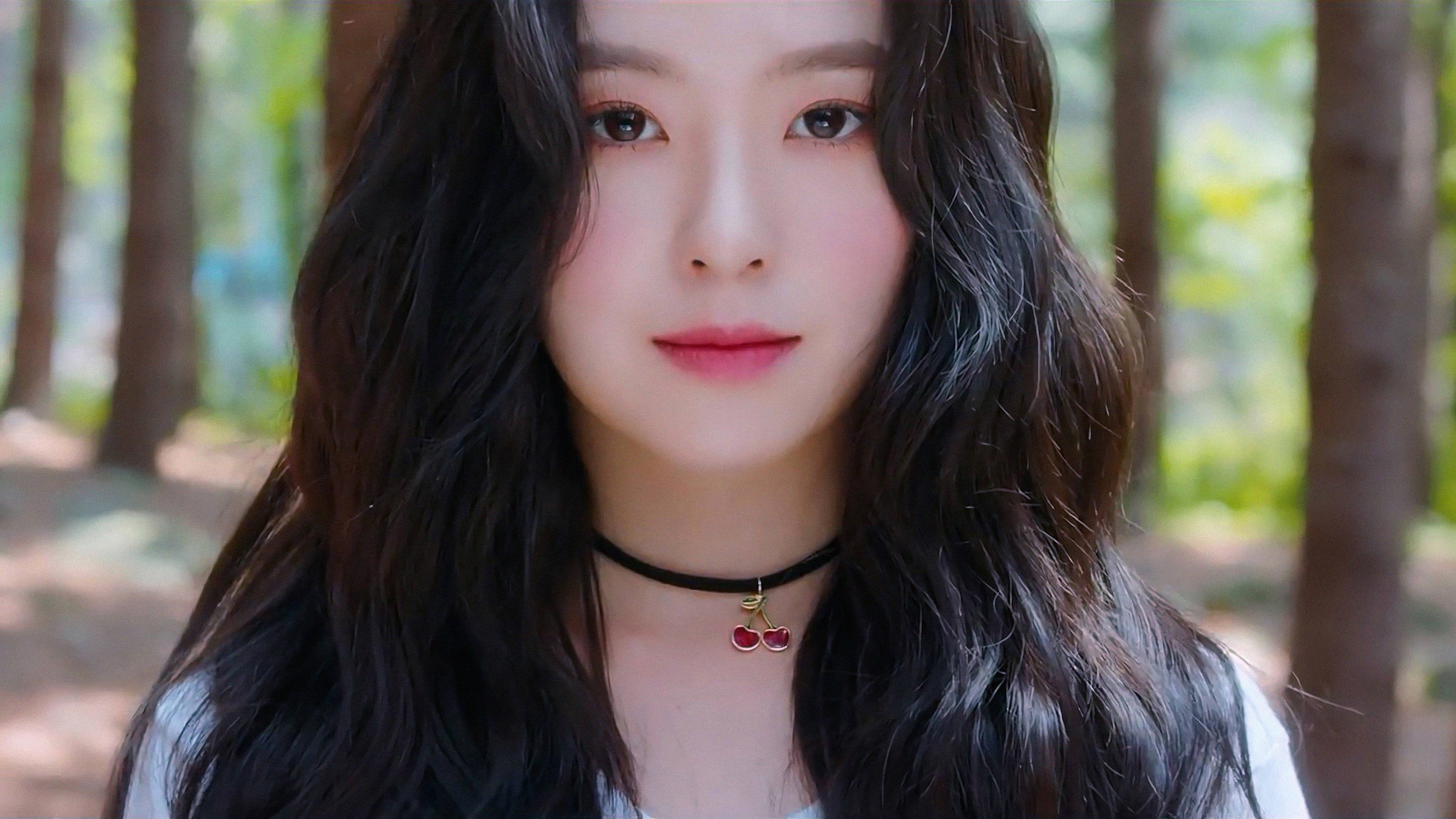 K-Pop Red Velvet Irene 壁紙 — 一位年輕女性，長著深色波浪長髮，戴著櫻桃吊墜頸鏈，在陽光明媚的森林中溫柔凝視。