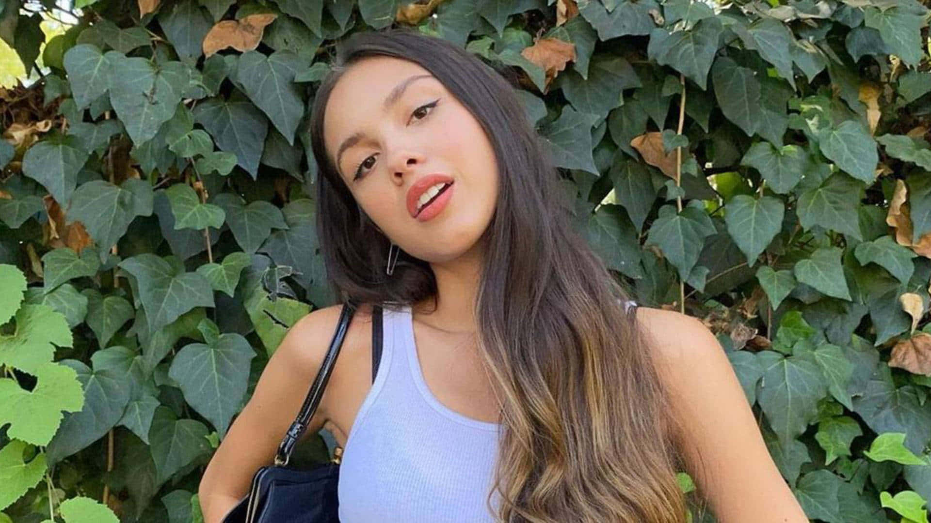 Olivia Rodrigo 桌布 壁紙 — 一位年輕女性，長著深色長髮，穿著淺藍色背心，自信地在茂密的綠色常春藤牆前擺姿勢。