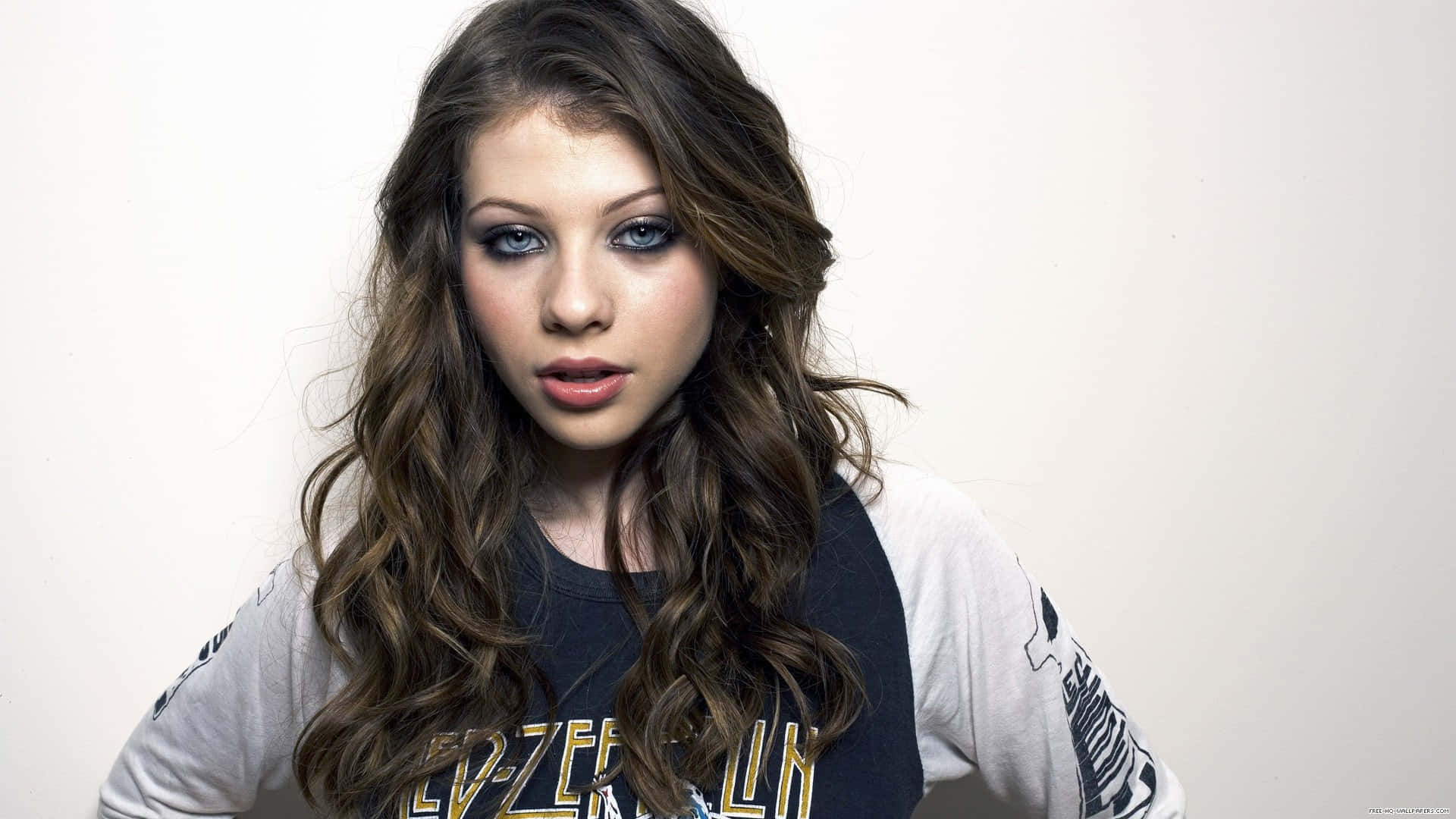 michelle trachtenberg 桌布 壁紙 — 一位年輕女性，長著長長的波浪棕色頭髮和大膽的藍色眼影，在純白色背景前直視鏡頭。