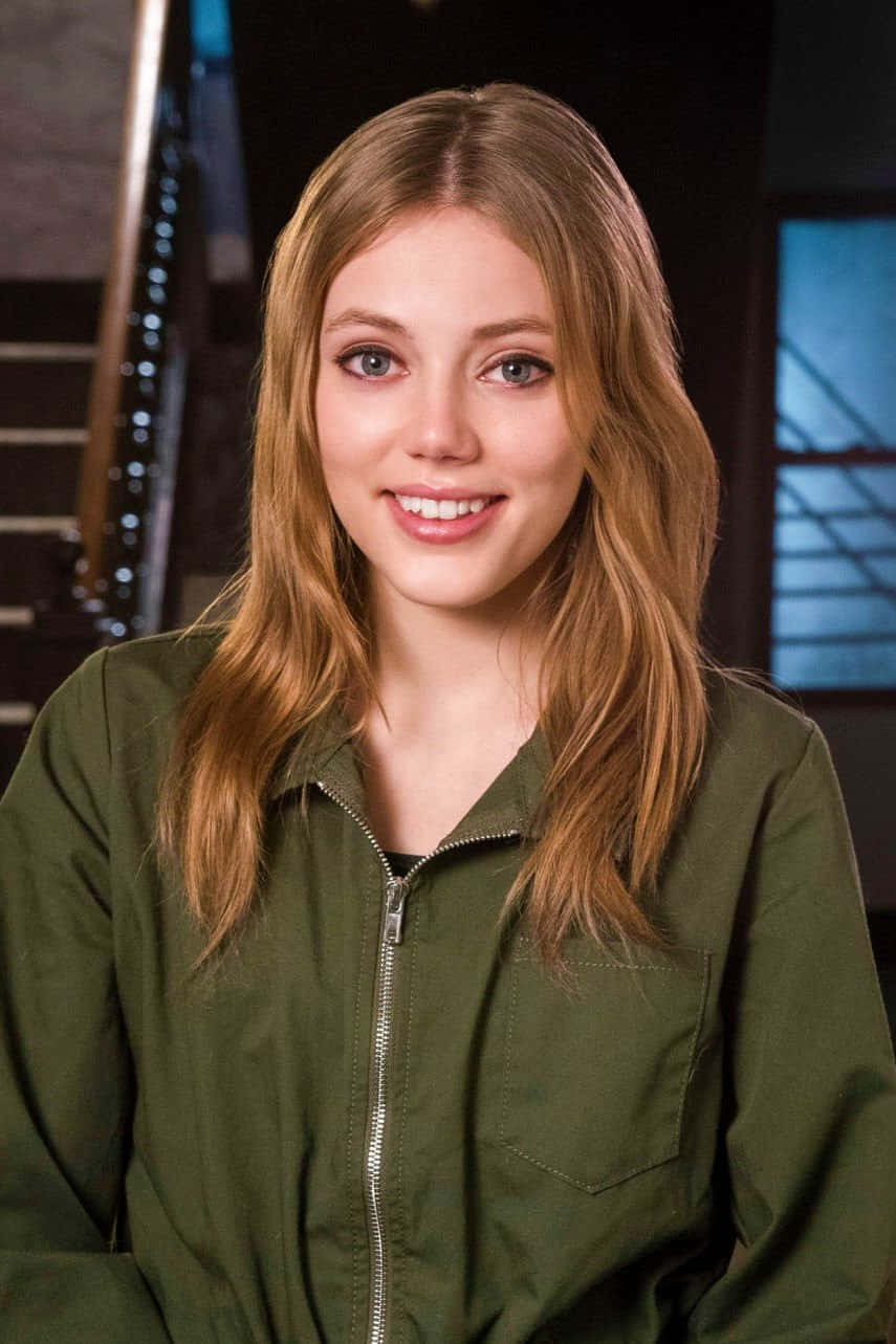 Grace Van Dien 桌布 壁紙 — 一位年輕女性，長長的金色頭髮，帶著溫暖的笑容，穿著橄欖綠拉鍊外套，背景是黑暗的城市環境，有樓梯和藍色燈光的窗戶。