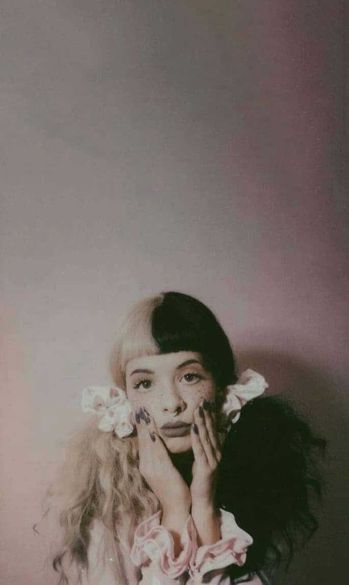 Melanie Martinez 桌布 壁紙 — 一位年輕女性，頭髮半黑半金，臉上有雀斑，雙手塗著深色指甲油，托著臉頰，背景是柔和的粉紅色。
