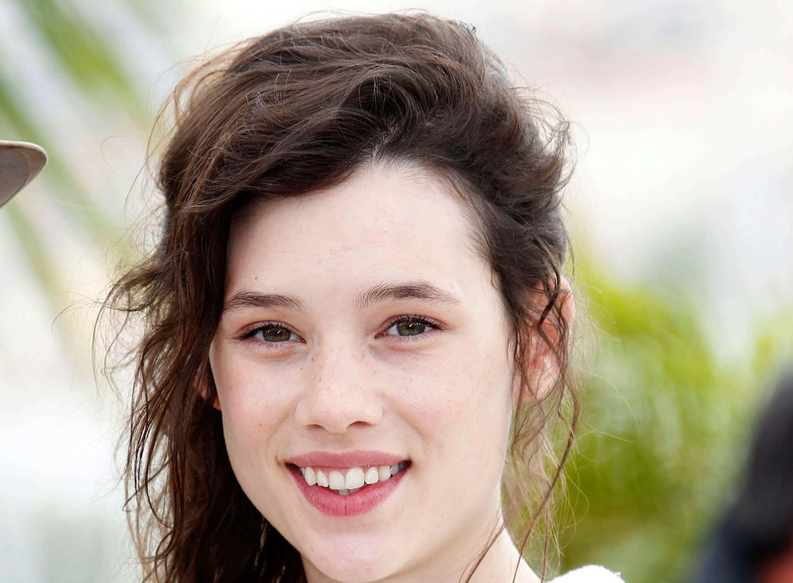 Astrid Berges Frisbey 桌布 壁紙 — 一位年輕女性嘅近距離肖像，佢有啲亂嘅棕色頭髮，喺柔和模糊嘅綠色背景下溫暖咁微笑。