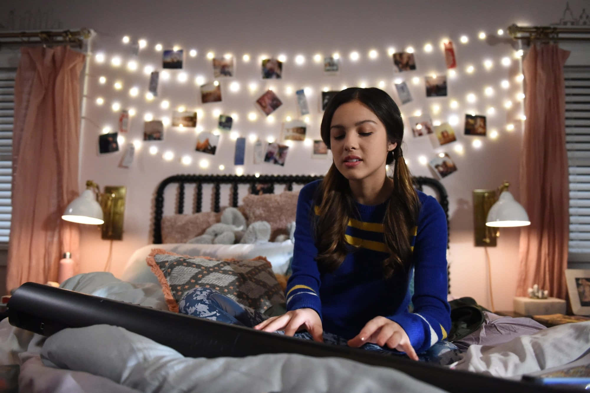 Olivia Rodrigo 桌布 壁紙 — 一位年輕女性坐在床上，周圍環繞著燈串和照片，在一個舒適、溫暖照明的臥室裡。