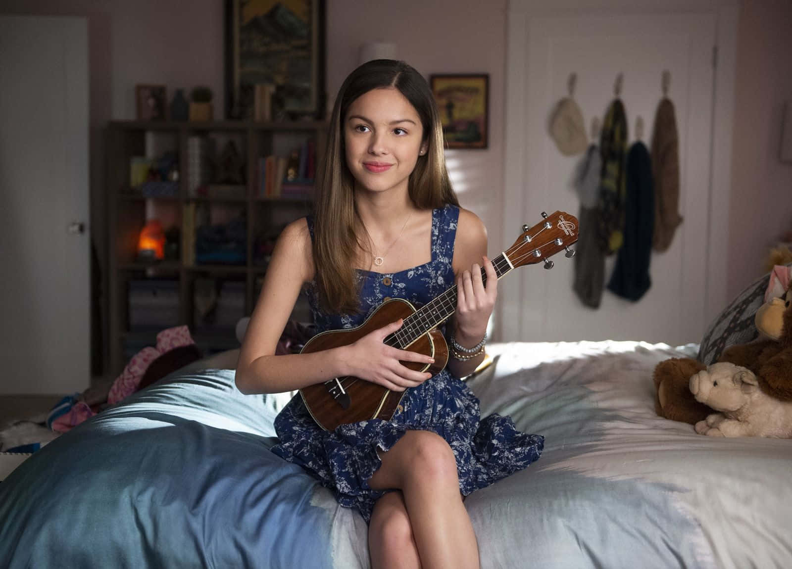 Olivia Rodrigo 桌布 壁紙 — 一位年輕女性坐在床上，在舒適的臥室裡拿著尤克里里，附近有柔和的燈光和毛絨玩具。