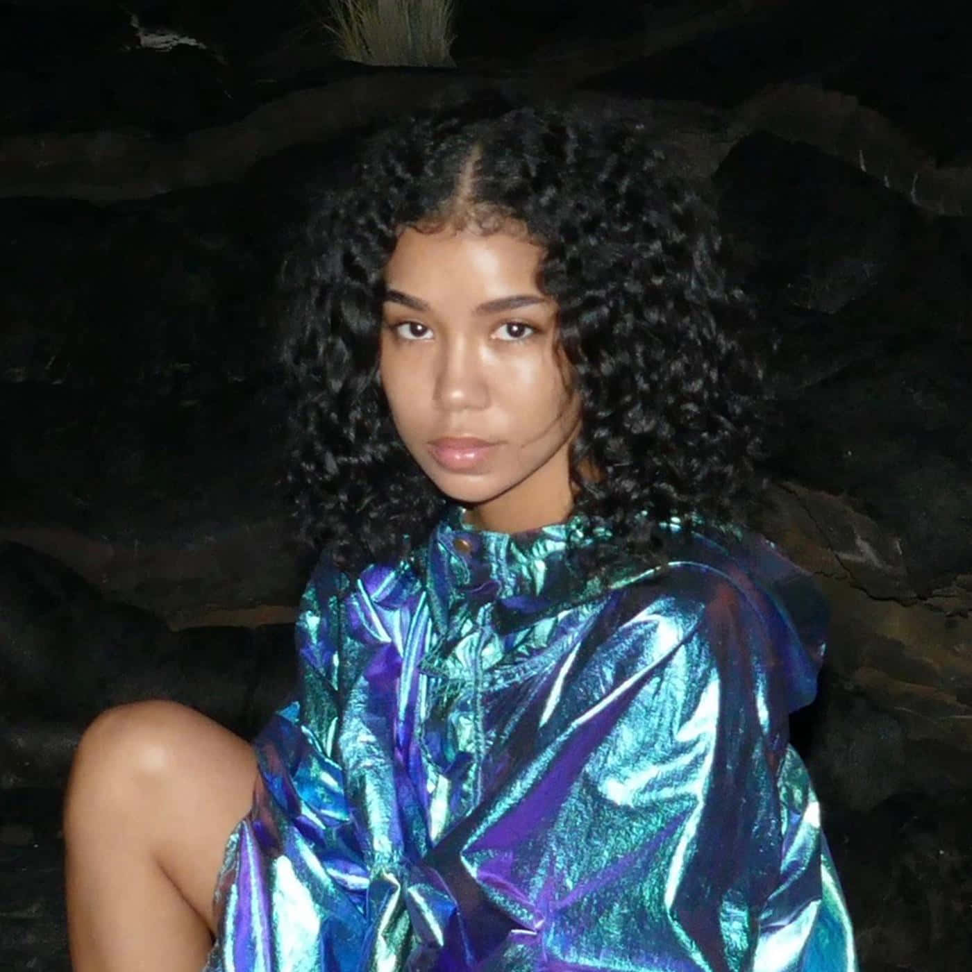 jhene aiko 桌布 壁紙 — 一位年輕女性擁有豐盈捲髮，靠著深色岩石，身穿閃爍虹彩藍紫色外套。