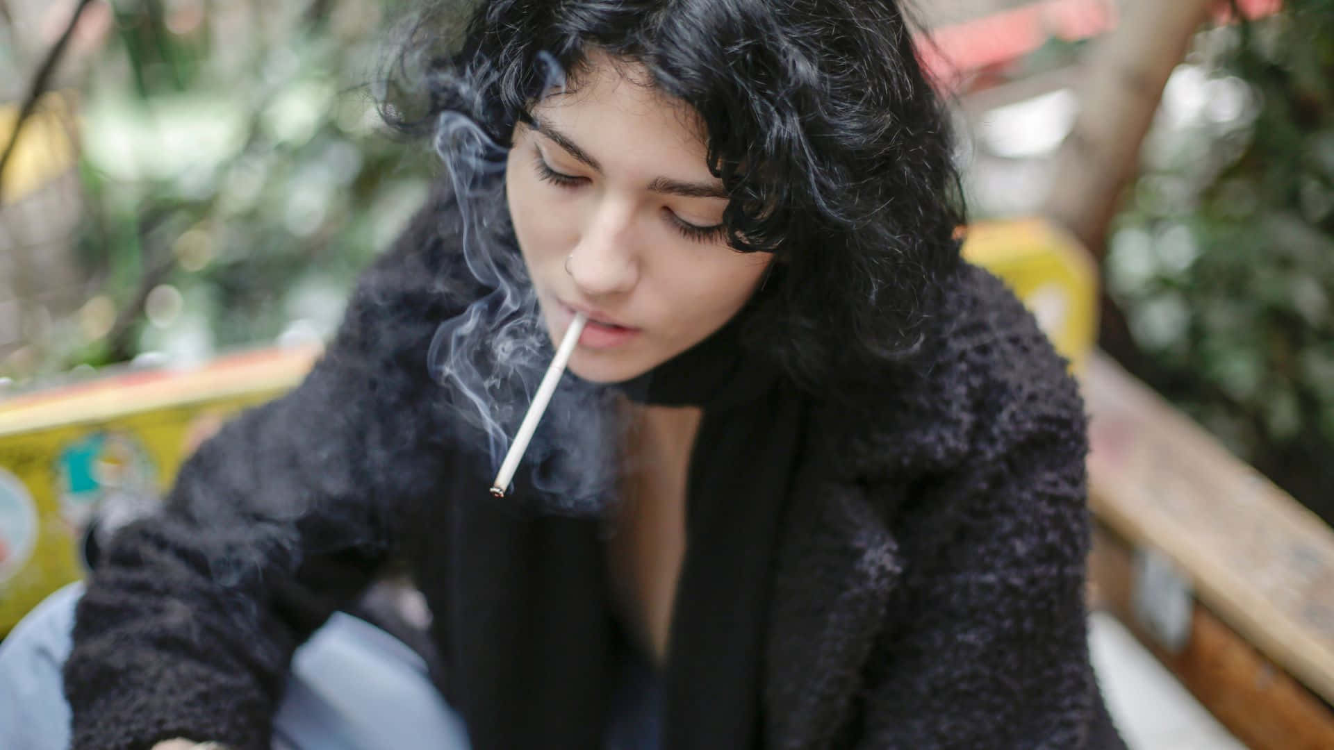 吸煙 桌布 壁紙 — 一位年輕女性留著深色捲髮，坐在戶外抽煙，煙霧在她臉龐縈繞。