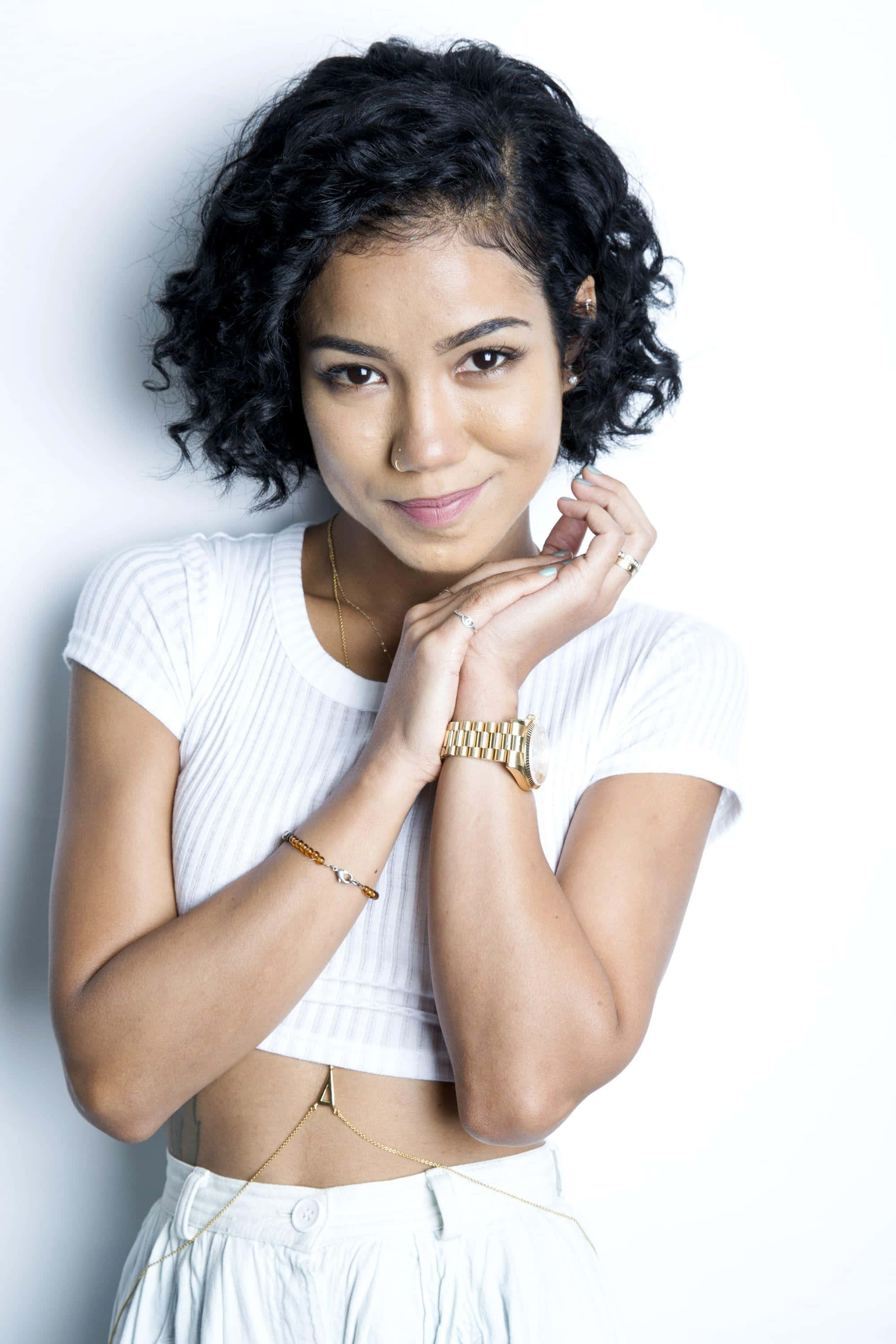 jhene aiko 桌布 壁紙 — 一位年輕女性留著黑色捲髮，身穿白色短版上衣和金色飾品，靠著白色牆壁。