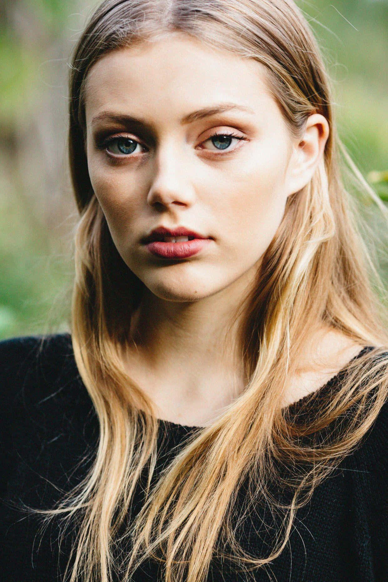 Grace Van Dien 桌布 壁紙 — 一位年輕女性的特寫肖像，擁有引人注目的藍色眼睛和金色頭髮，穿著黑色上衣，背景是柔和的綠色模糊背景。