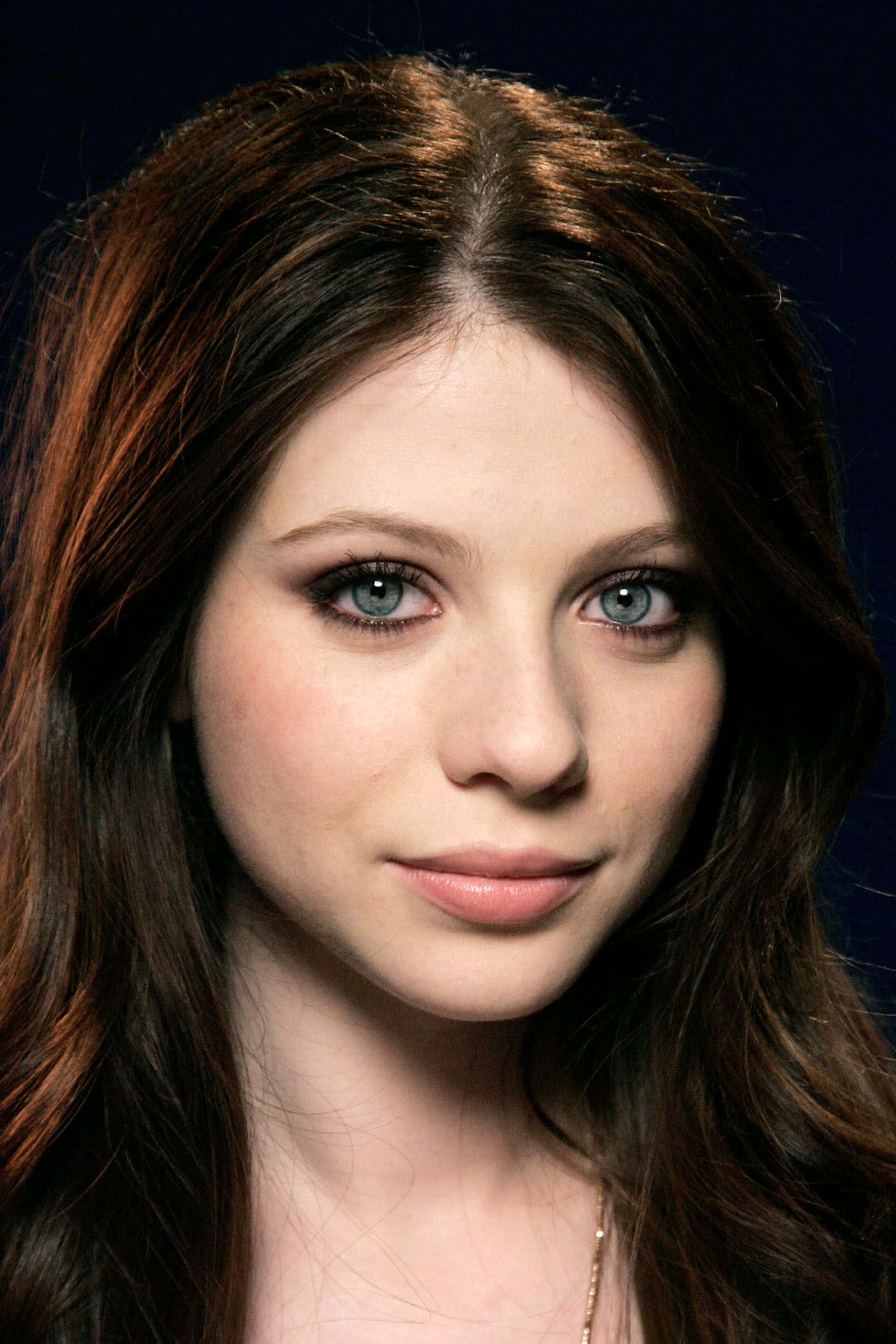 michelle trachtenberg 桌布 壁紙 — 一位年輕女性的特寫肖像，擁有引人注目的藍色眼睛和長長的棕色頭髮，背景為深藍色。
