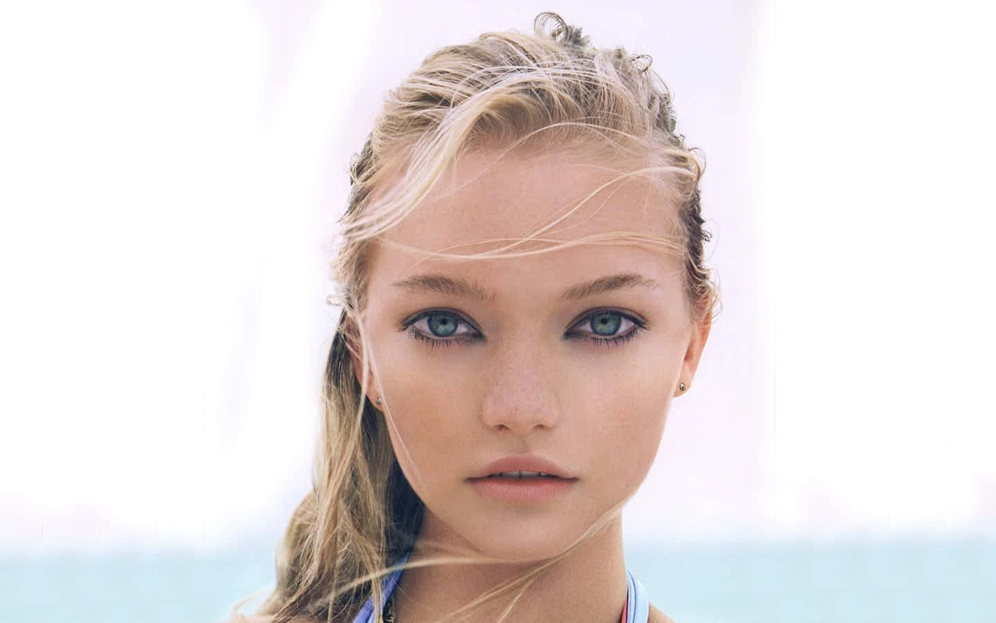 Gemma Ward 桌布 壁紙 — 一位年輕女性的特寫肖像，擁有引人注目的藍色眼睛和隨風飄揚的金色頭髮，背景明亮而模糊。