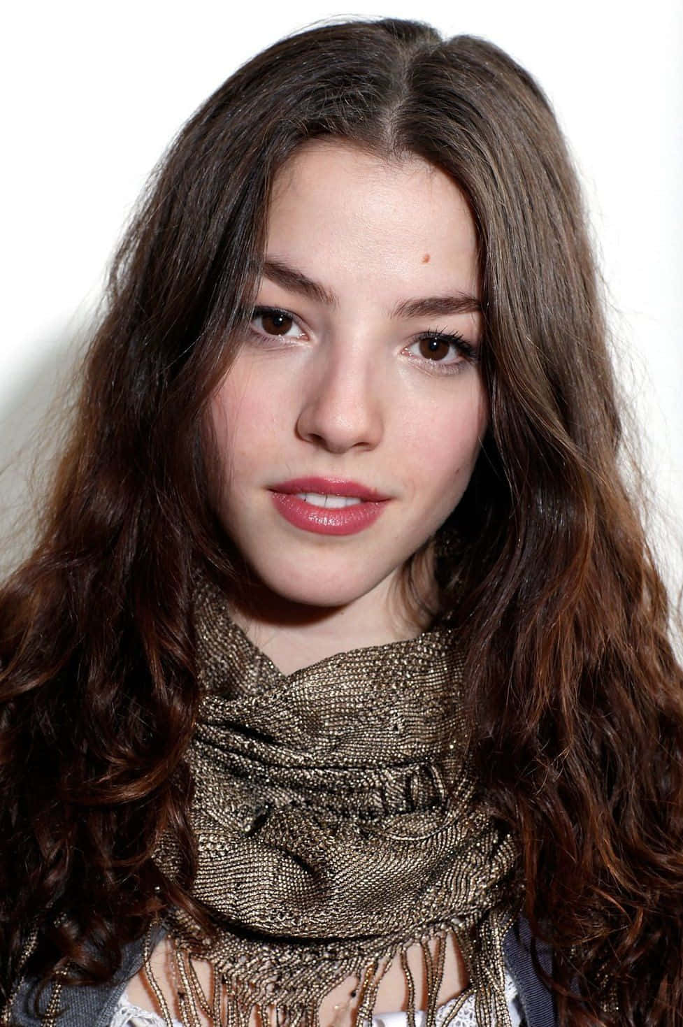 Olivia Thirlby 桌布 壁紙 — 一位年輕女性的特寫肖像，擁有長長的波浪形棕色頭髮和一條閃閃發光的金色圍巾，背景是純白色。