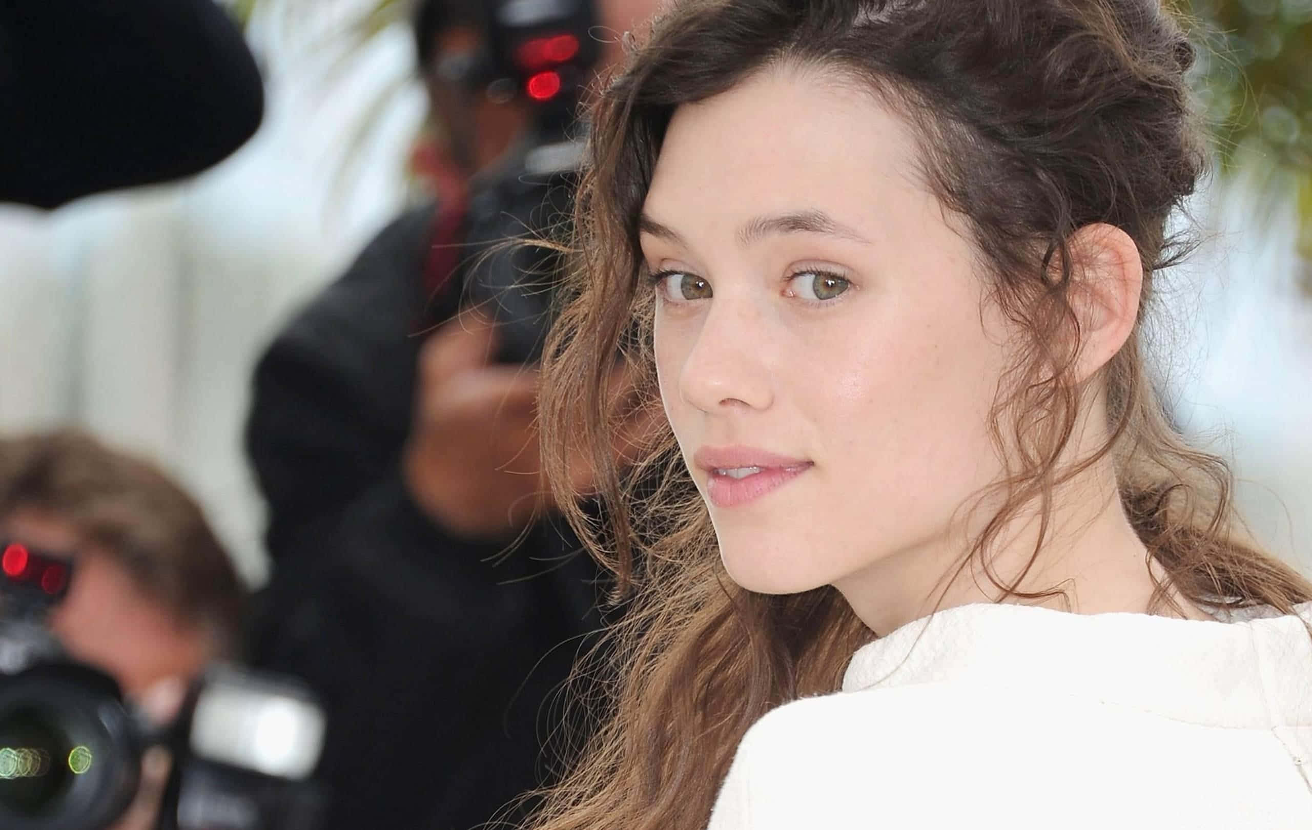 Astrid Berges Frisbey 桌布 壁紙 — 一位年輕女性的特寫肖像，棕色頭髮略顯凌亂，眼神柔和，身穿白色上衣，周圍是模糊的攝影師和紅色相機燈光。