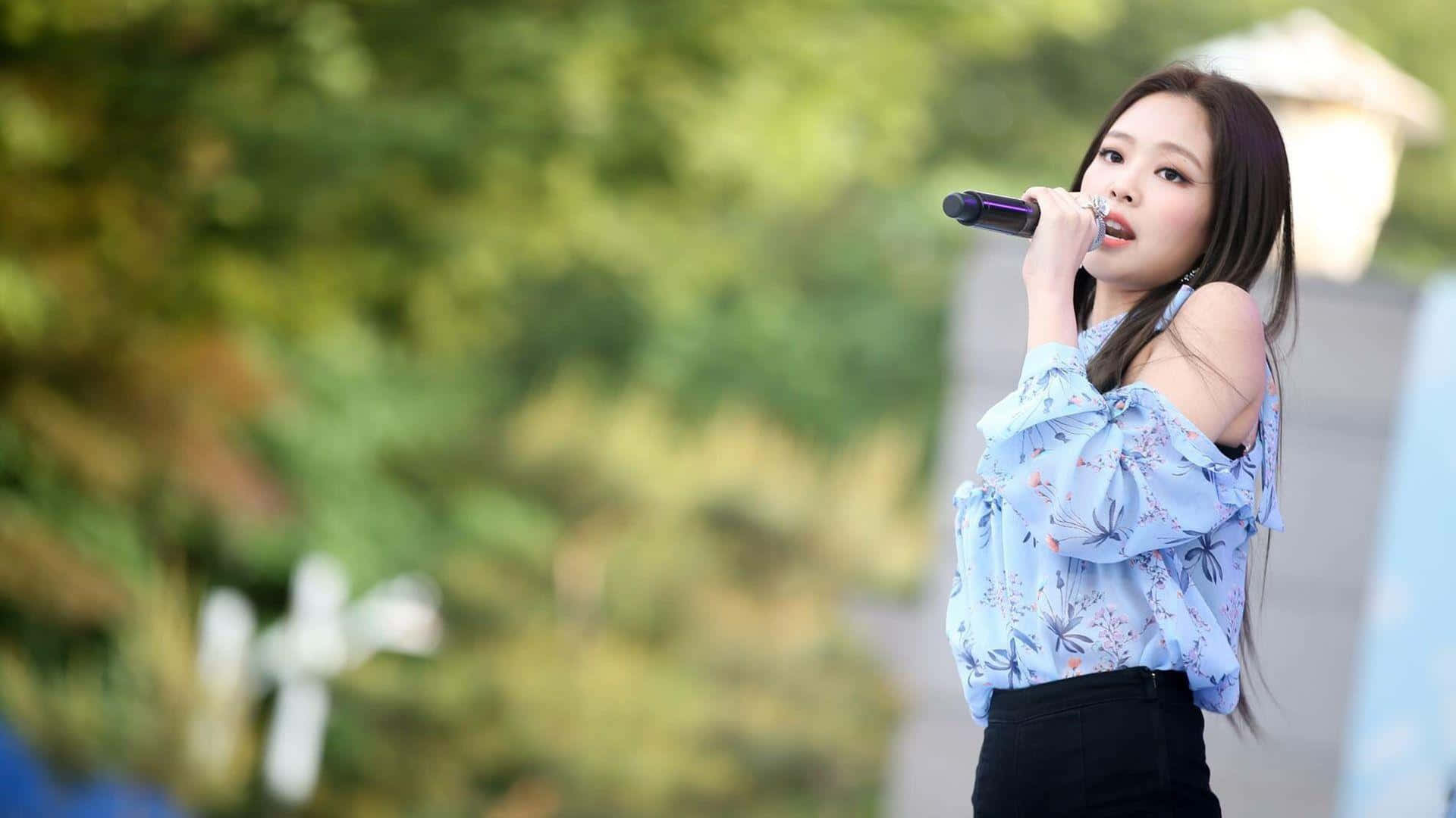 Jennie Kim 桌布 壁紙 — 一位年輕女性穿著花卉圖案藍色襯衫，在模糊的綠色戶外背景下，熱情地對著麥克風唱歌。
