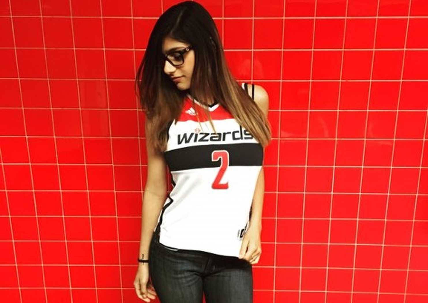 Mia Khalifa 桌布 壁紙 — 一位年輕女性身穿巫師隊籃球球衣，靠著大膽的紅色瓷磚牆，戴著眼鏡，低頭看著下方。