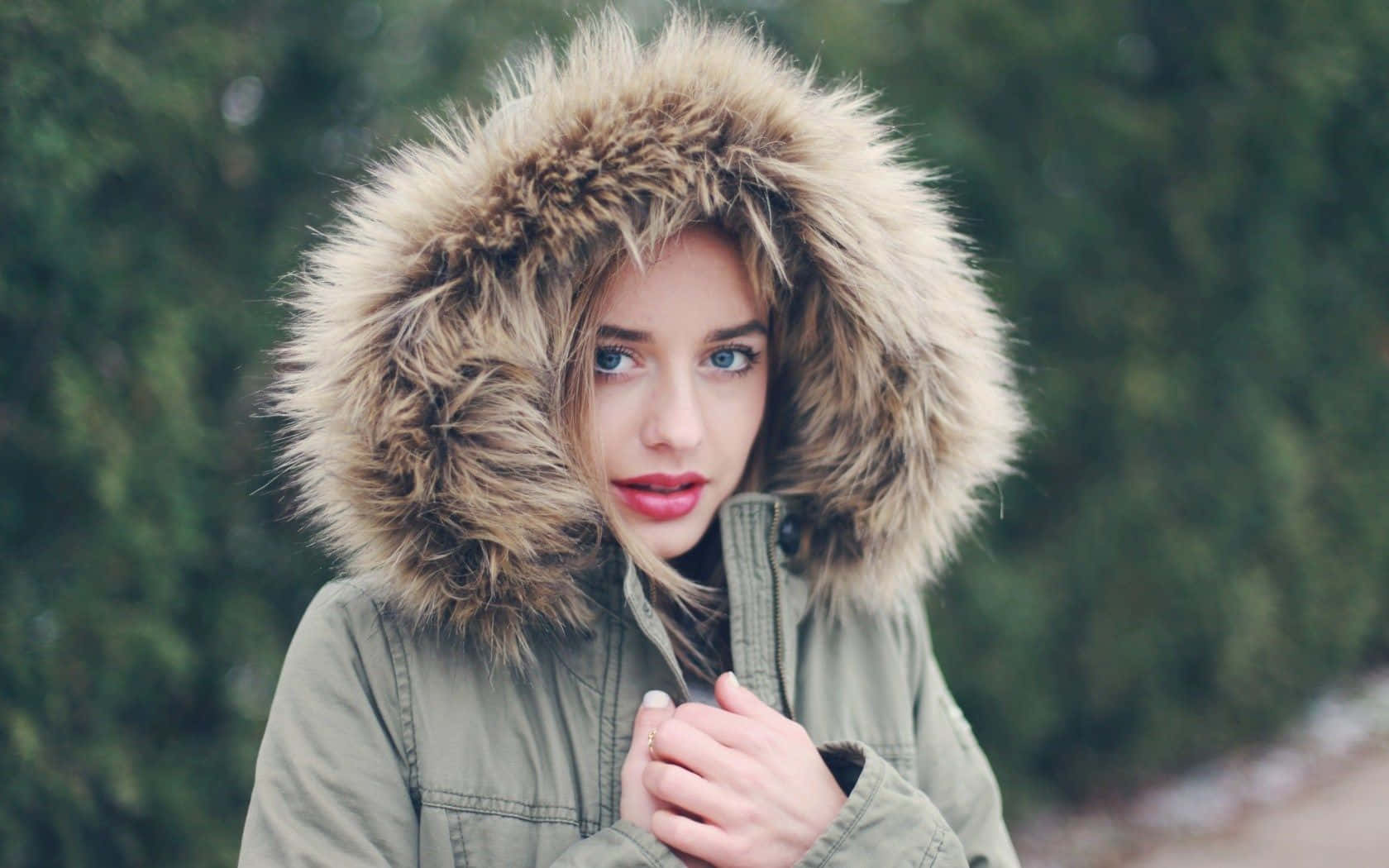 Winter Fashion 桌布 壁紙 — 一位年輕女性身穿綠色派克大衣，戴著毛茸茸的連帽，在模糊的森林背景下直視鏡頭。