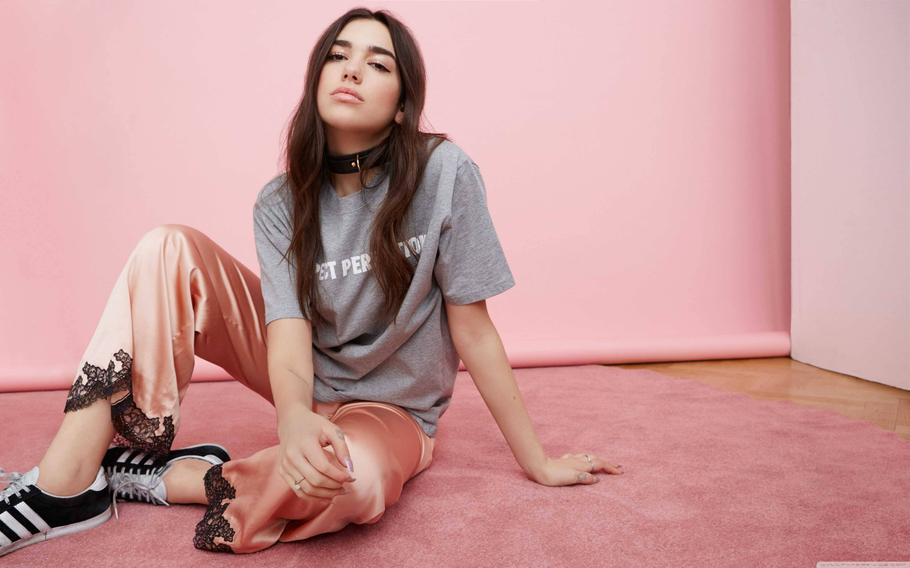Dua Lipa 4K Spotify Sessions 壁紙 — 一位年輕女性隨意地坐在粉紅色地毯上，背景是相配的粉紅色牆壁，身穿灰色T恤、桃色緞面褲子和黑色運動鞋。