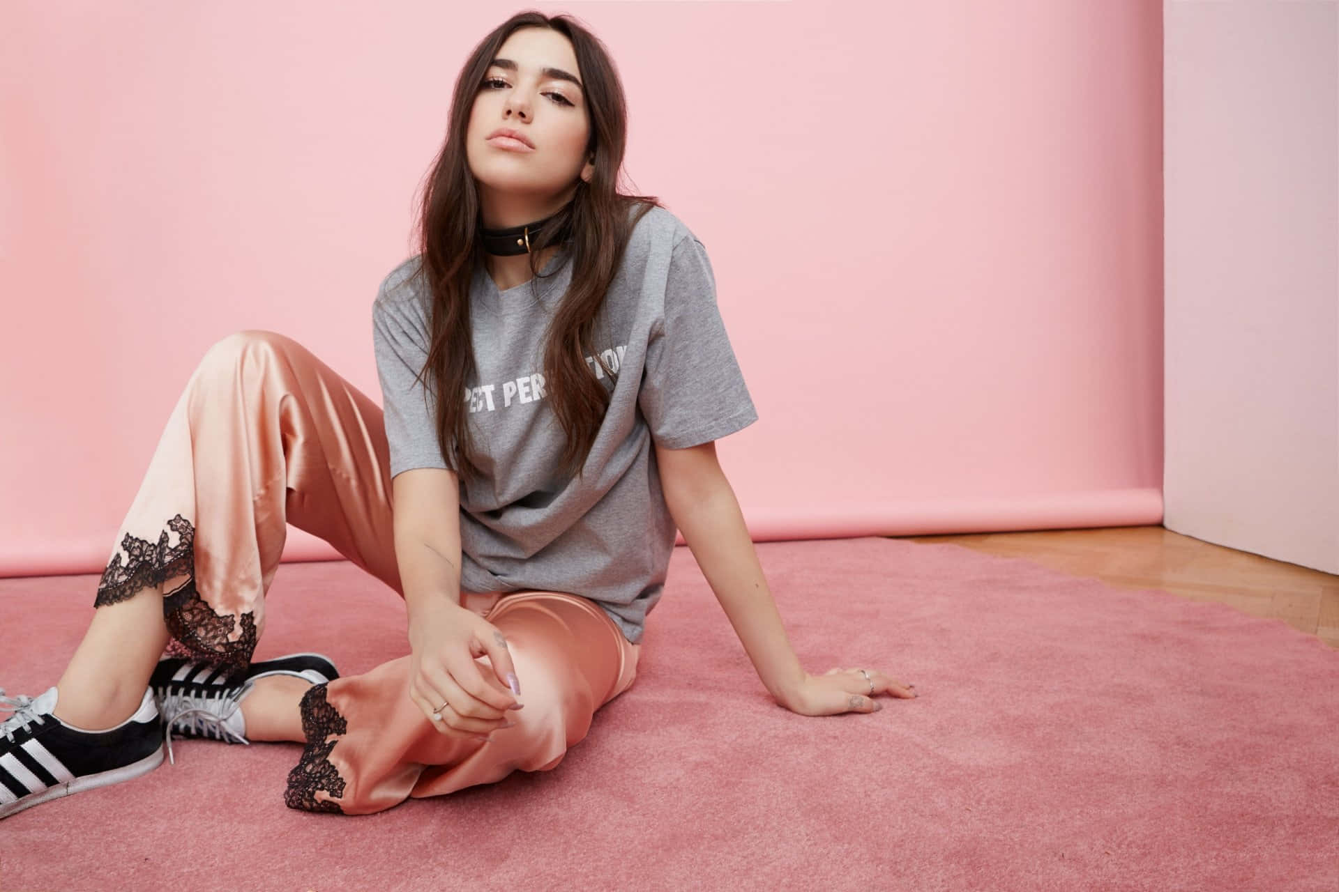 Dua Lipa 桌布 壁紙 — 一位年輕女性隨意地坐在粉紅色地毯上，背景是相配的粉紅色牆壁，身穿灰色T恤、桃色緞面褲子和黑色運動鞋。