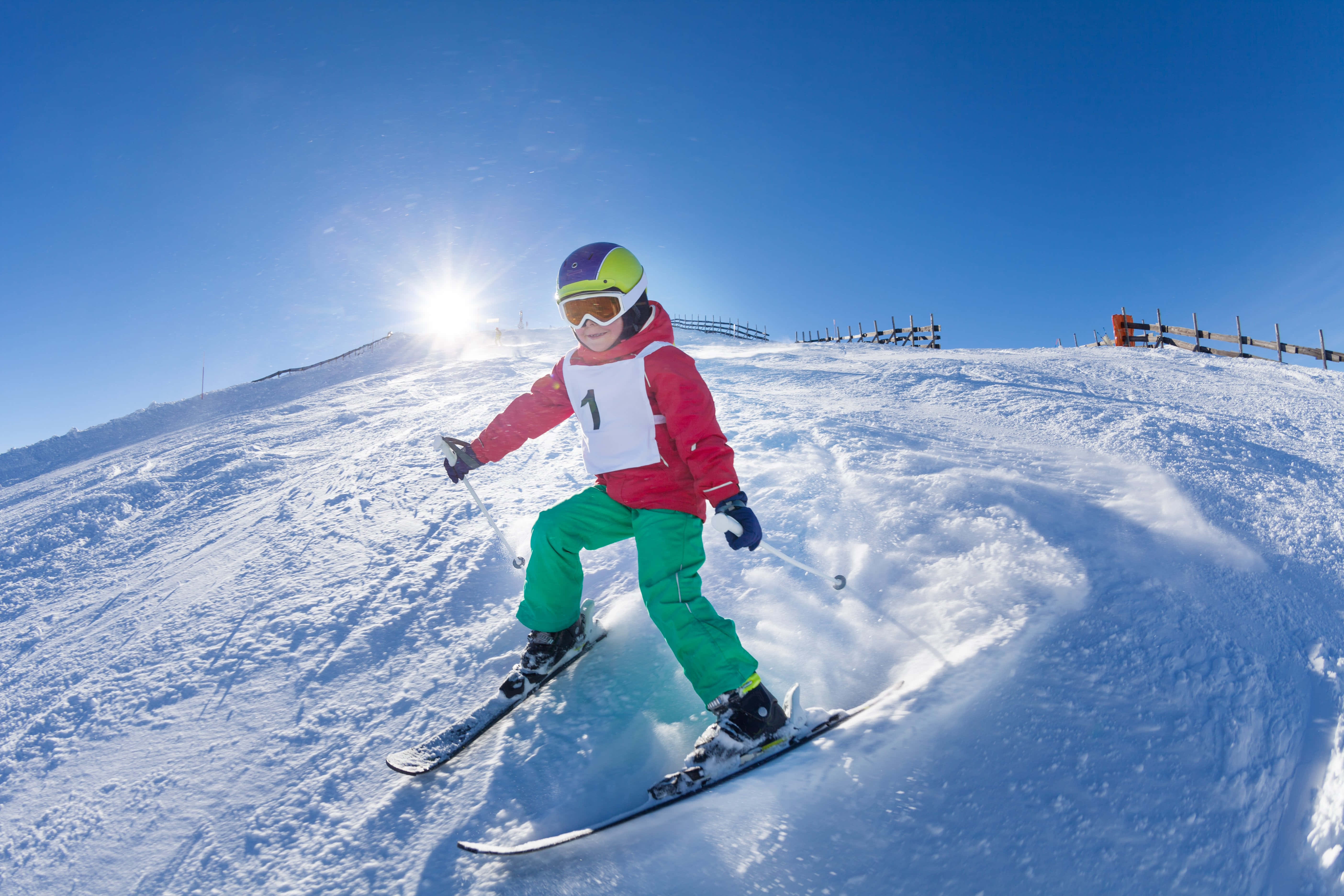 Skiing 桌布 壁紙 — 一位年輕滑雪者穿著鮮豔裝備，在晴朗藍天下陽光普照的雪坡上劃過新鮮的粉雪。