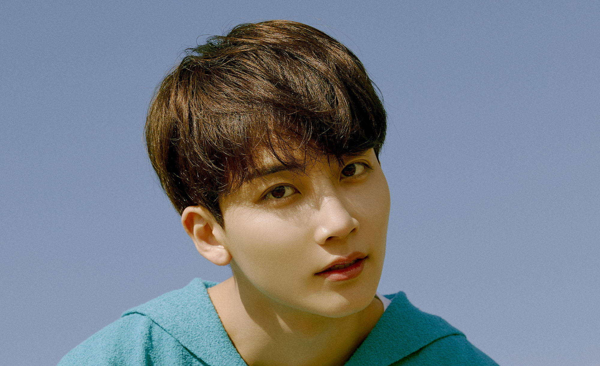 Yoon Jeonghan Henggarae 壁紙 — 一位年輕男子，一頭凌亂的棕色頭髮，穿著青綠色連帽衫，在清澈的藍天下若有所思地凝視著。
