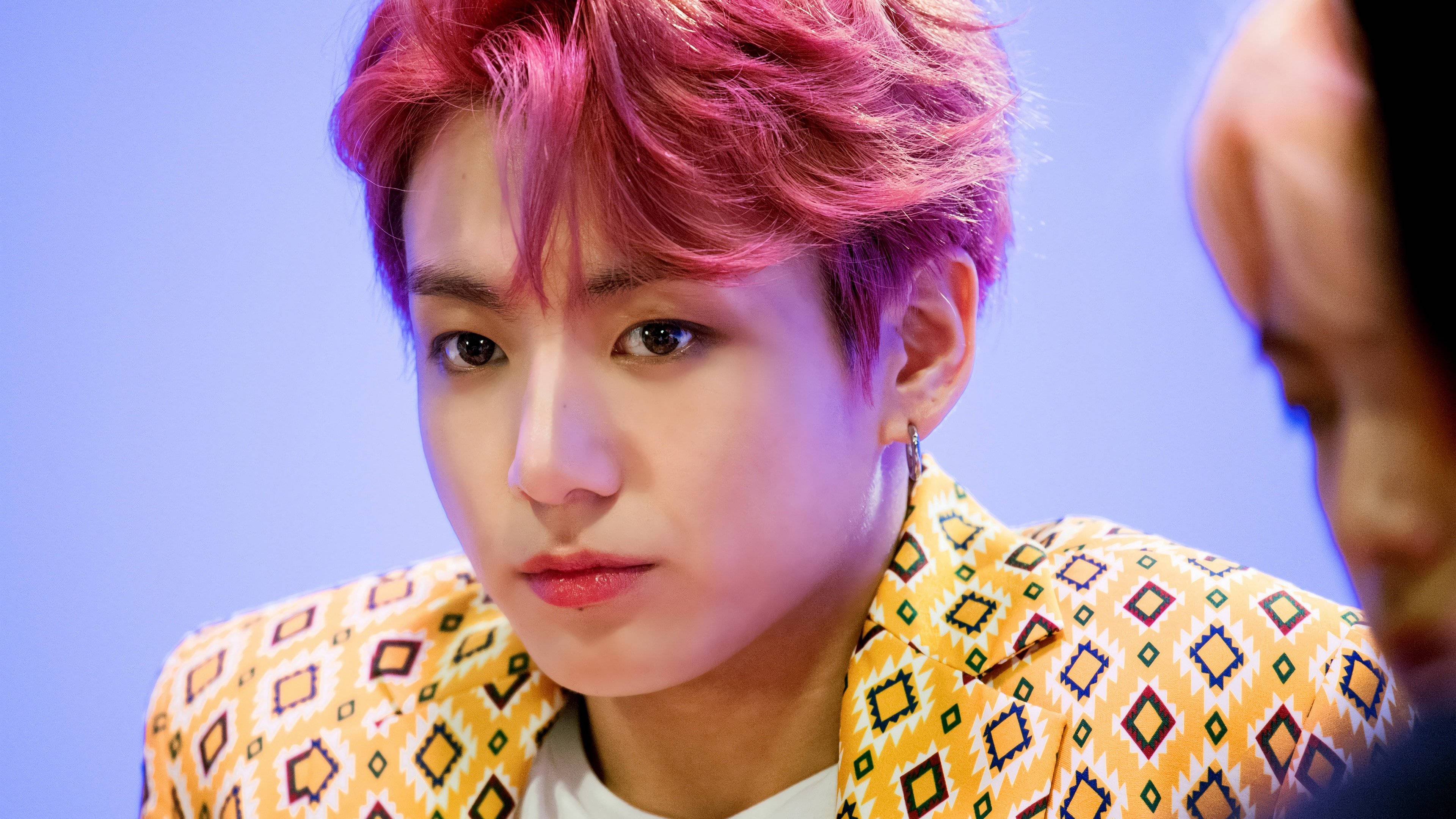 Idol Jeon Jungkook 壁紙 — 一位年輕男子，擁有鮮豔的粉紅色頭髮和圖案黃色外套，在柔和的藍色背景下若有所思地凝視。