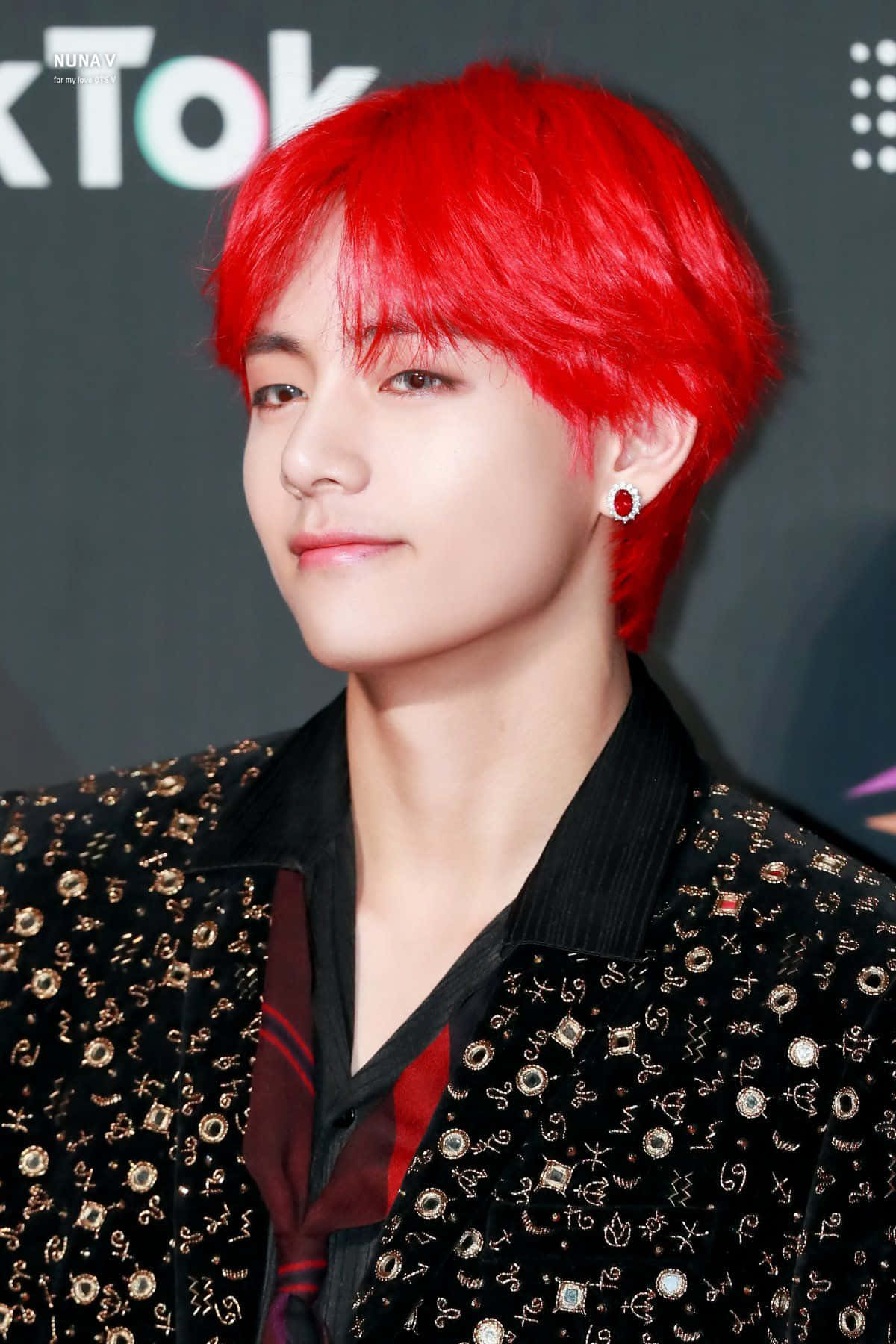 Taehyung Cute 桌布 壁紙 — 一位年輕男子，擁有鮮豔的紅髮和紅色寶石耳環，身穿飾有金色符號的黑色外套，背景為深色。