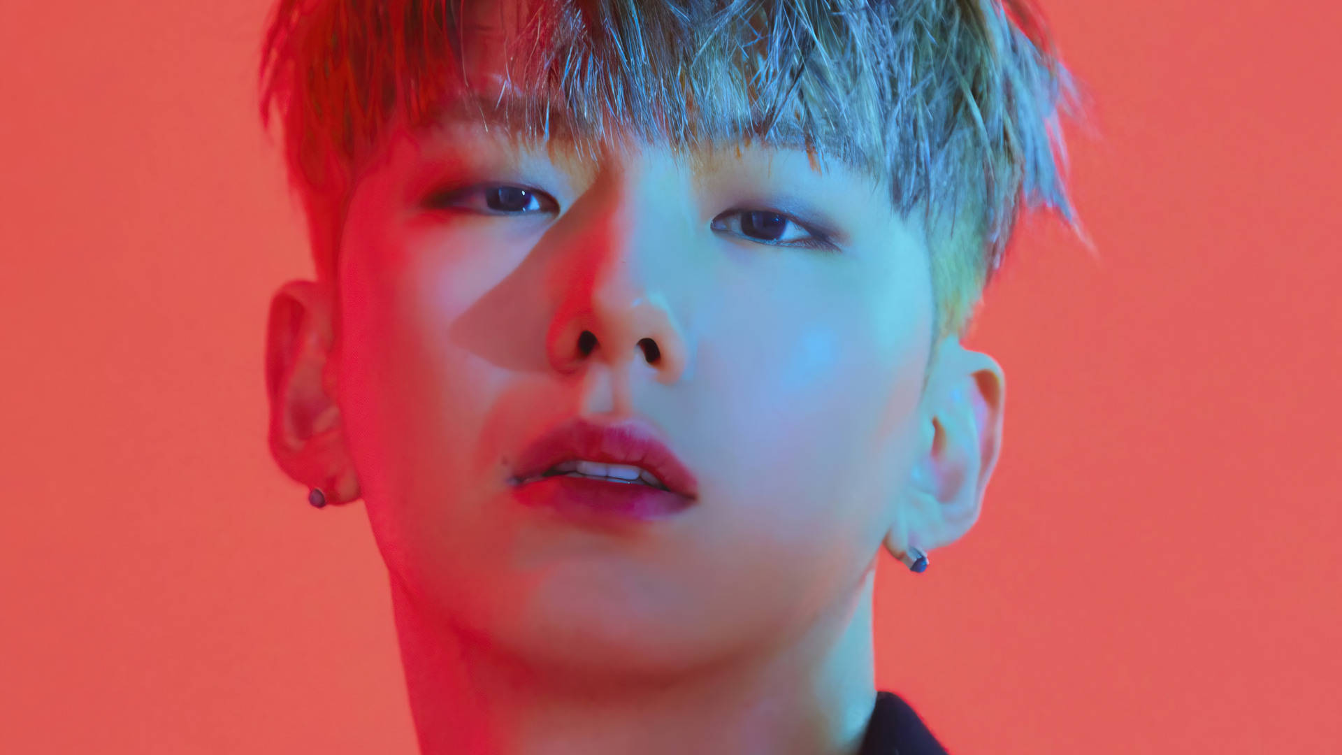 Monsta X Kihyun Fatal Love 壁紙 — 一位年輕男子，梳著時尚髮型並戴著耳環，在戲劇性的紅藍燈光下，以純橙色背景為襯，凝視著鏡頭。