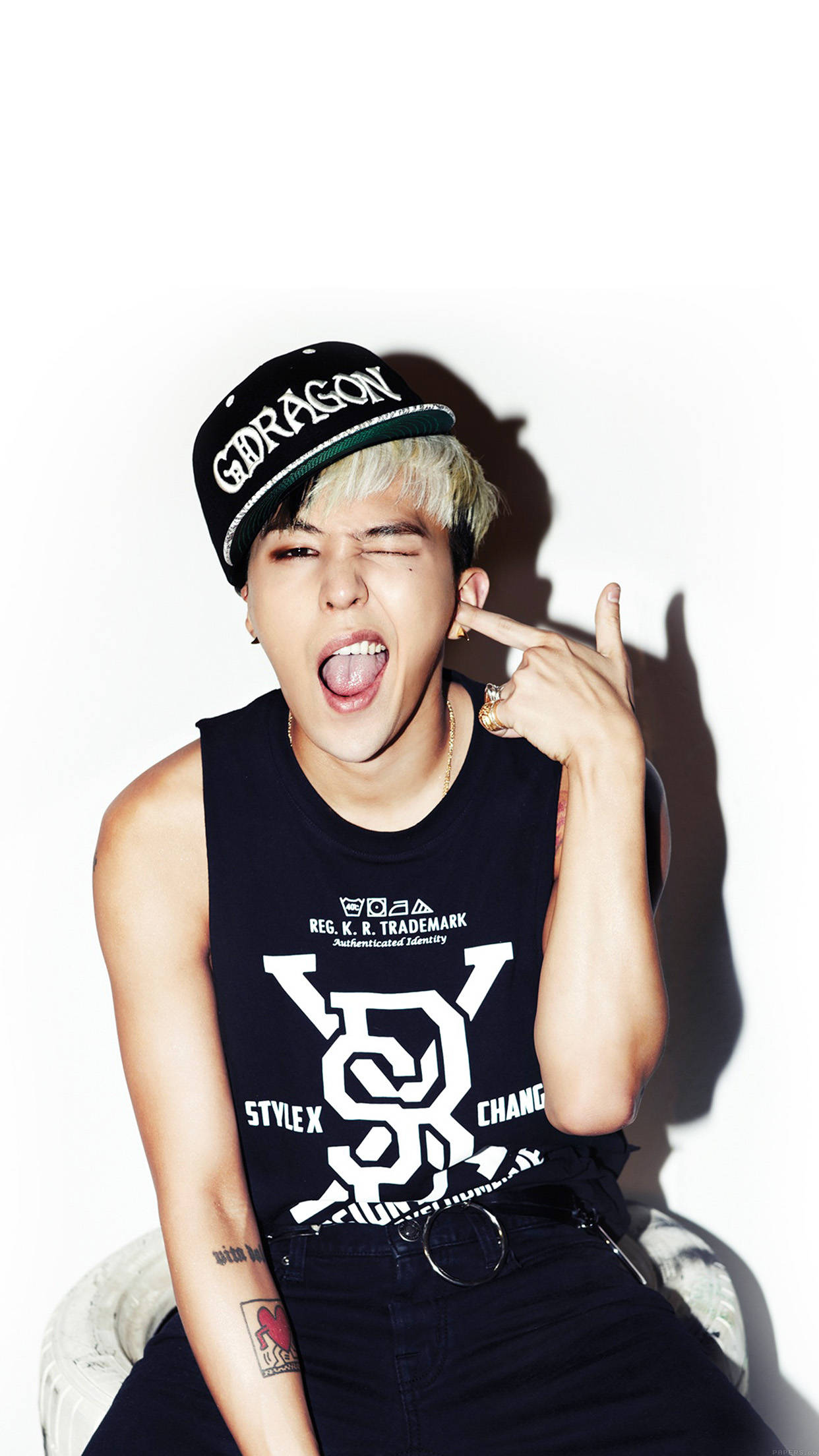 G-dragon 《Crooked》 壁紙 — 一位年輕男子，漂染頭髮，戴著黑色鴨舌帽，在純白背景前眨眼並伸出舌頭，同時做出手勢。
