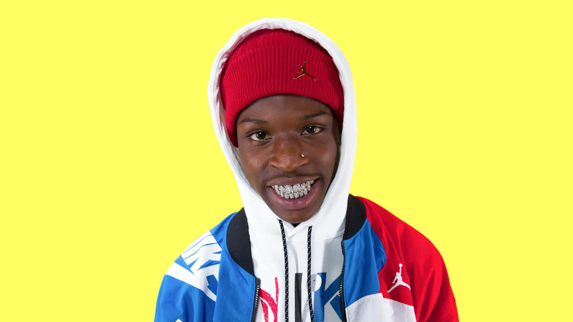 Quando Rondo 桌布 壁紙 — 一位年輕男子帶著燦爛笑容和鑽石牙套，戴著紅色冷帽，穿著色彩繽紛的Jordan連帽衫，背景是鮮黃色。