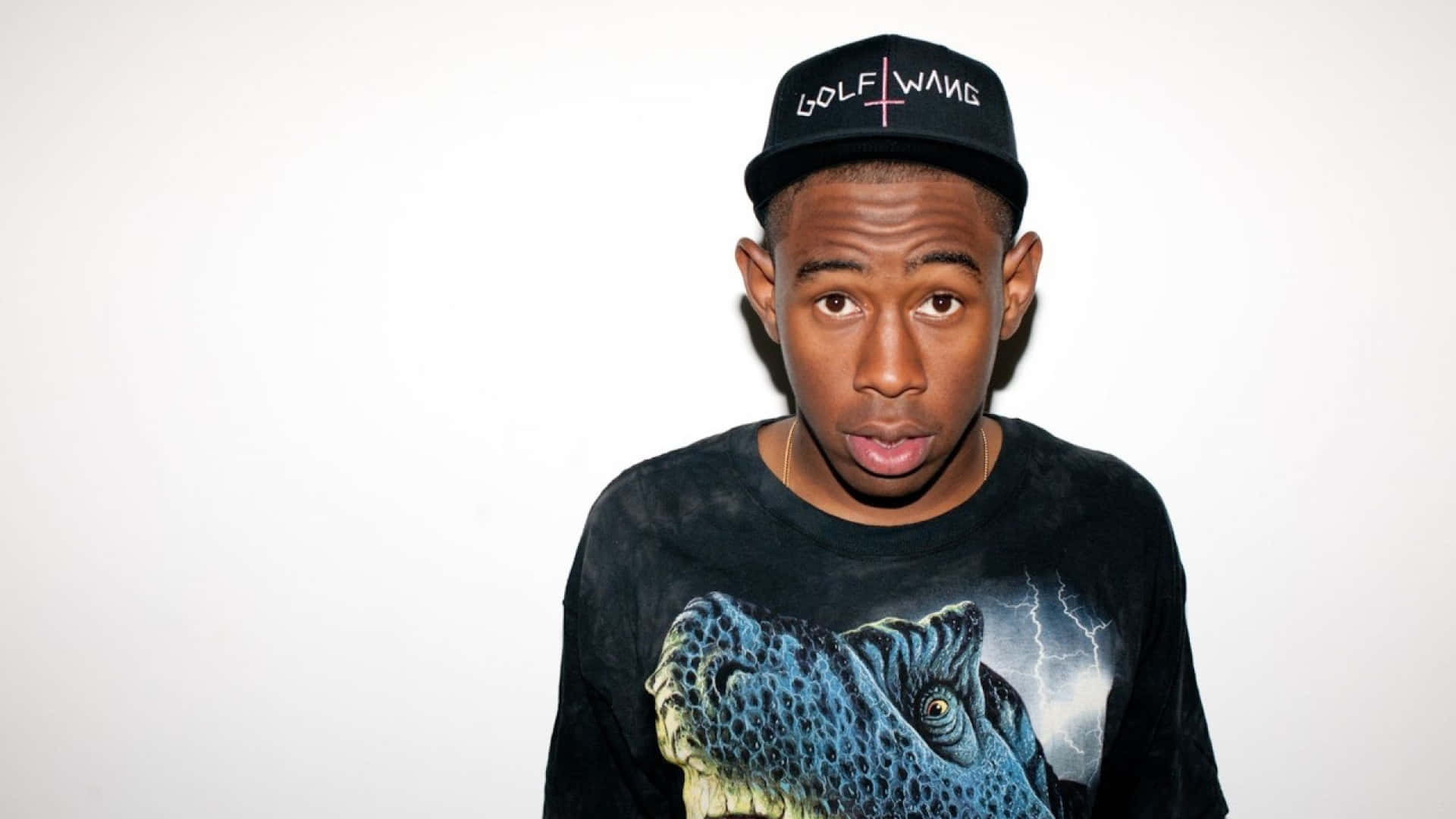 tyler the creator 桌布 壁紙 — 一位年輕男子戴著黑色Golf Wang帽，身穿恐龍印花T恤，在純白背景下直視鏡頭。