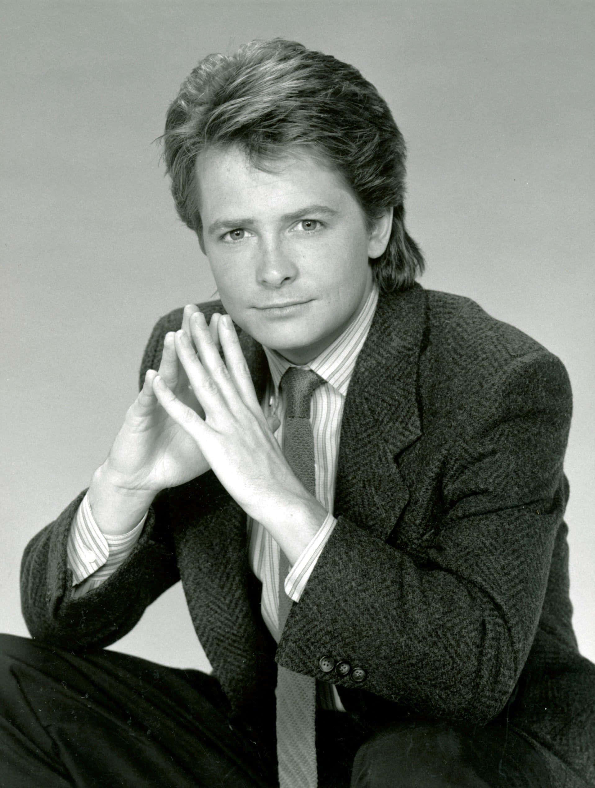 michael j fox 桌布 壁紙 — 一位年輕男子梳著時尚髮型，穿著質感西裝外套和條紋襯衫，雙手交握靠近臉部，坐在攝影棚內。