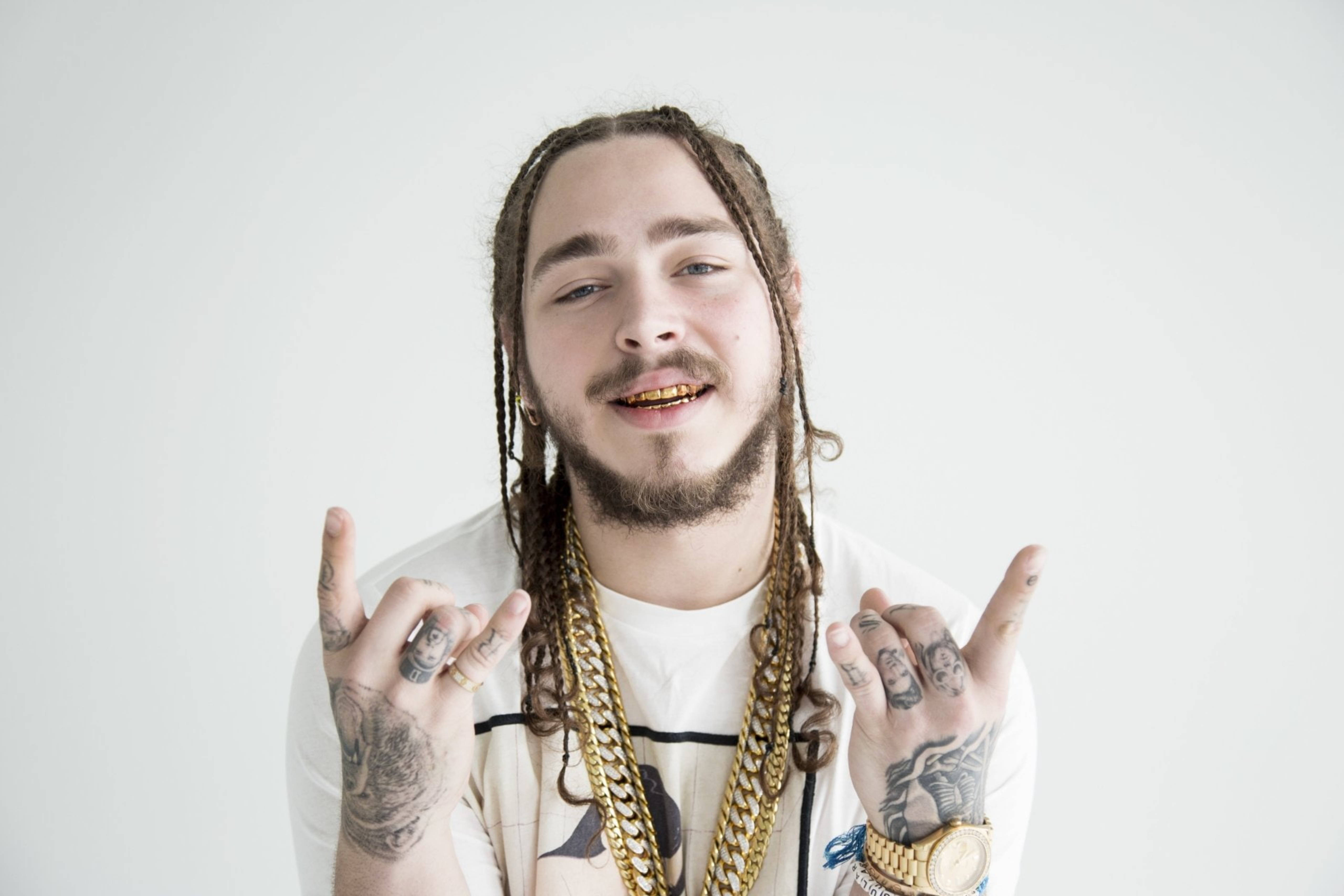 Post Malone GQ 2016 壁紙 — 一位年輕男子編著辮子，戴著金牙套，在純白背景前微笑並做出搖滾手勢。
