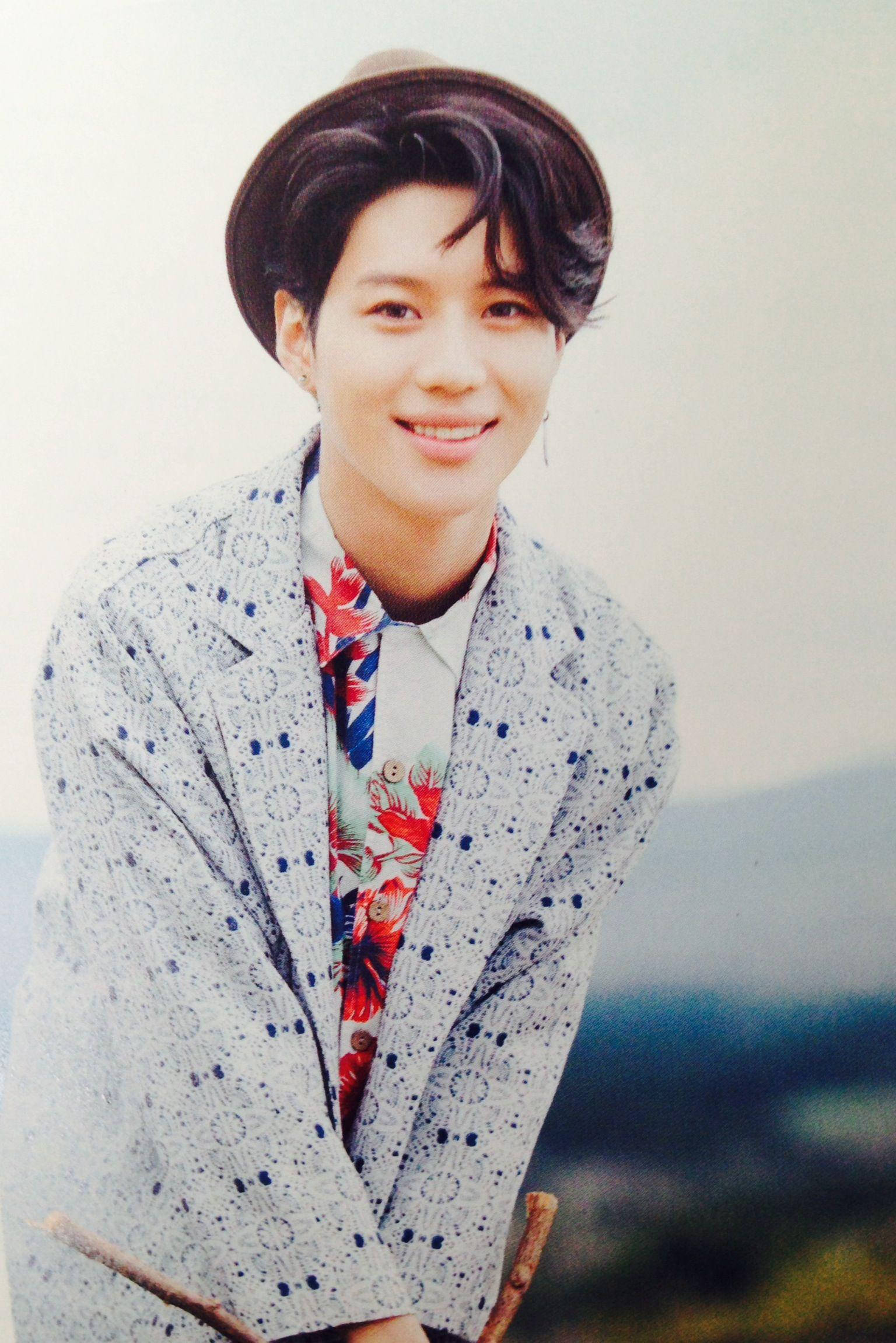 Shinee Young Taemin 壁紙 — 一位年輕男子身穿圖案西裝外套和花卉襯衫，戴著棕色帽子，在模糊的戶外背景下溫暖地微笑。