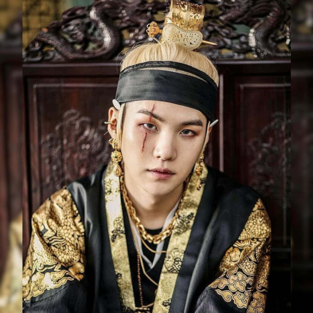 Suga 桌布 壁紙 — 一位年輕男子身穿華麗的黑金傳統韓國王室服飾，帶有龍紋圖案和一隻血紅的眼睛，在雕刻木質背景前凝視著。