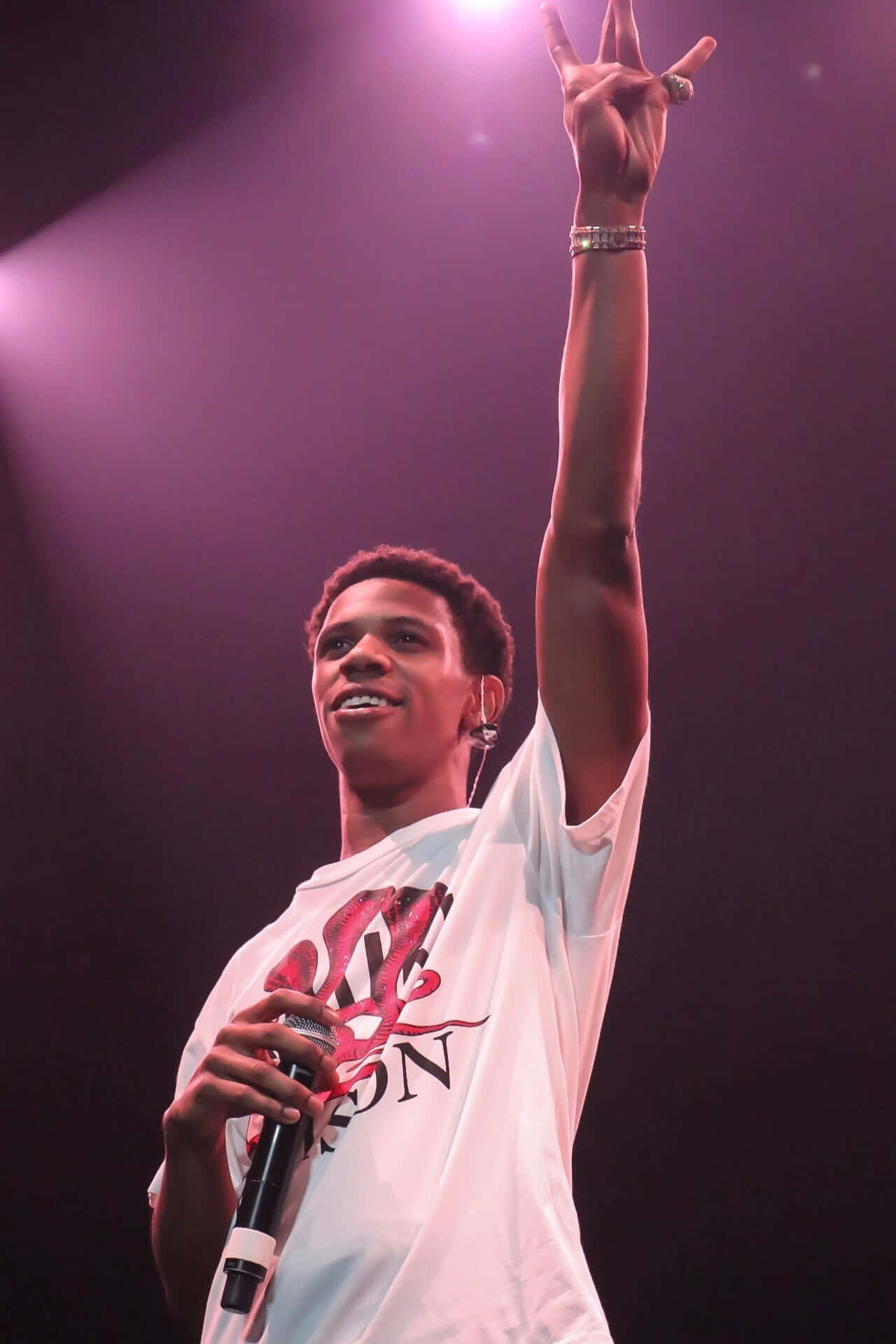 A Boogie Wit Da Hoodie 壁紙 — 一位年輕男表演者在紫色聚光燈下燦爛微笑，手持麥克風，舉起手做出和平手勢。
