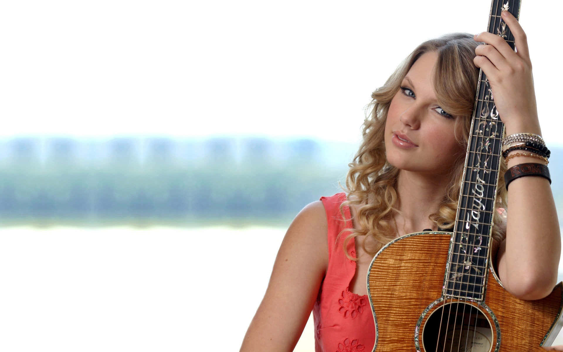 Taylor Swift 桌布 壁紙 — 一位年輕的捲曲金髮女子在明亮模糊的戶外背景下抱著一把原聲結他。