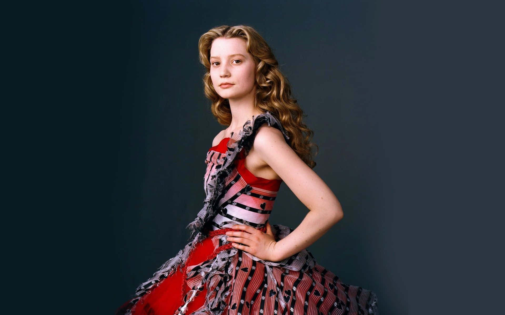 Mia Wasikowska 桌布 壁紙 — 一位年輕的捲曲金髮女性，身穿紅色圖案連衣裙，在深藍綠色工作室背景前自信地擺姿勢。