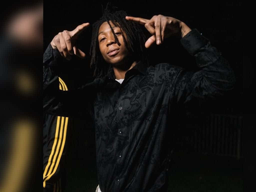 Dead Rappers Man 壁紙 — 一位年輕的辮子男在昏暗的肖像照中指向自己的頭部，身穿帶有黃色條紋的黑色圖案外套。