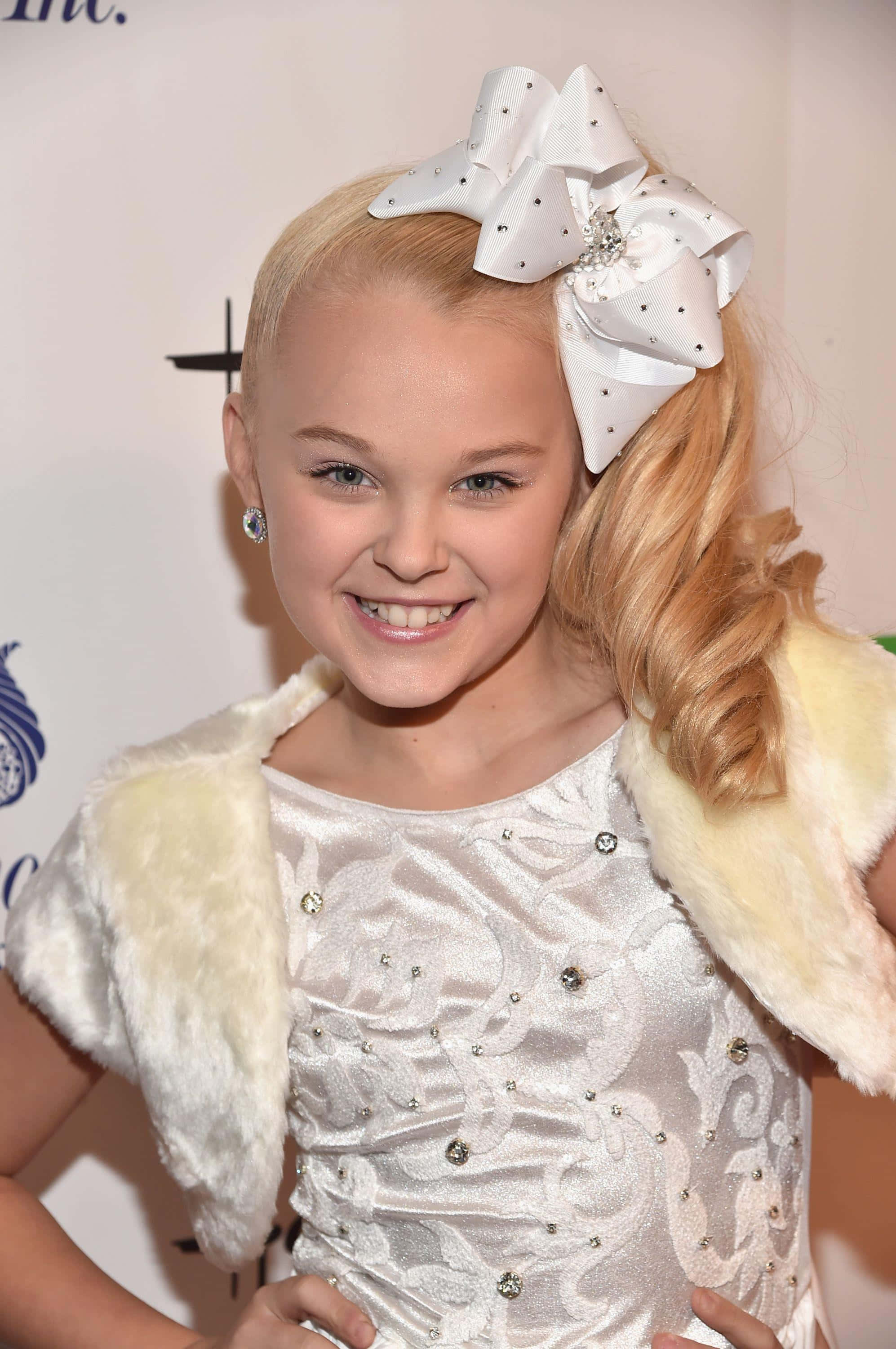 jojo siwa 桌布 壁紙 — 一位年輕的金髮女孩穿著白色亮片連衣裙，頭上戴著一個大大的白色蝴蝶結，露出燦爛的笑容。