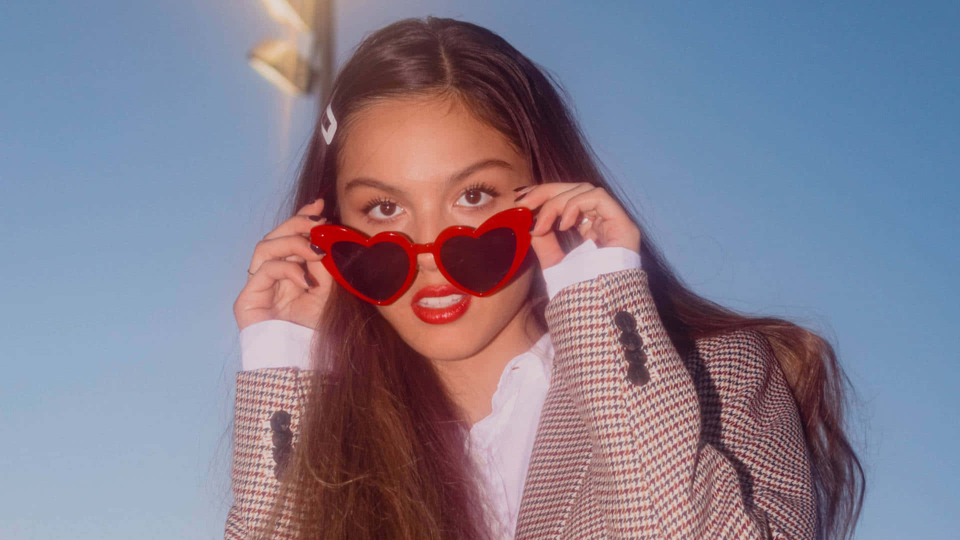 Olivia Rodrigo 桌布 壁紙 — 一位年輕的長髮棕髮女性戴著大膽的紅色心形太陽眼鏡，背景是清澈的藍天。