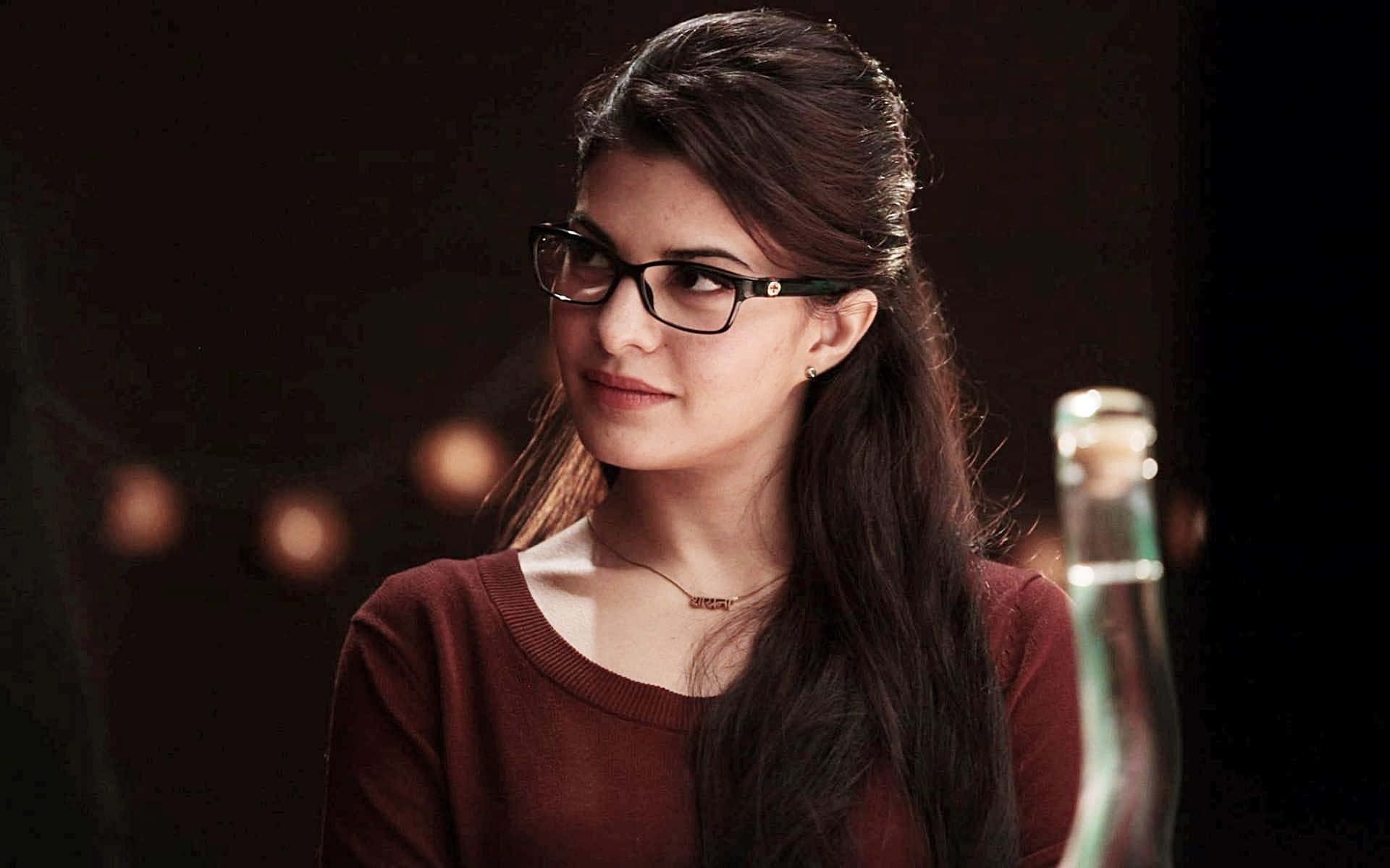 Jacqueline Fernandez 桌布 壁紙 — 一位年輕的黑髮女子戴著眼鏡，在昏暗的房間裡若有所思地凝視，背景是溫暖的散景燈光。