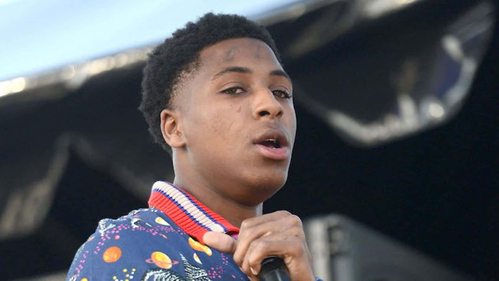 NBA YoungBoy 桌布 壁紙 — 一位年輕黑人男表演者在明亮的日光下，穿著色彩繽紛的太空主題襯衫對著麥克風唱歌。
