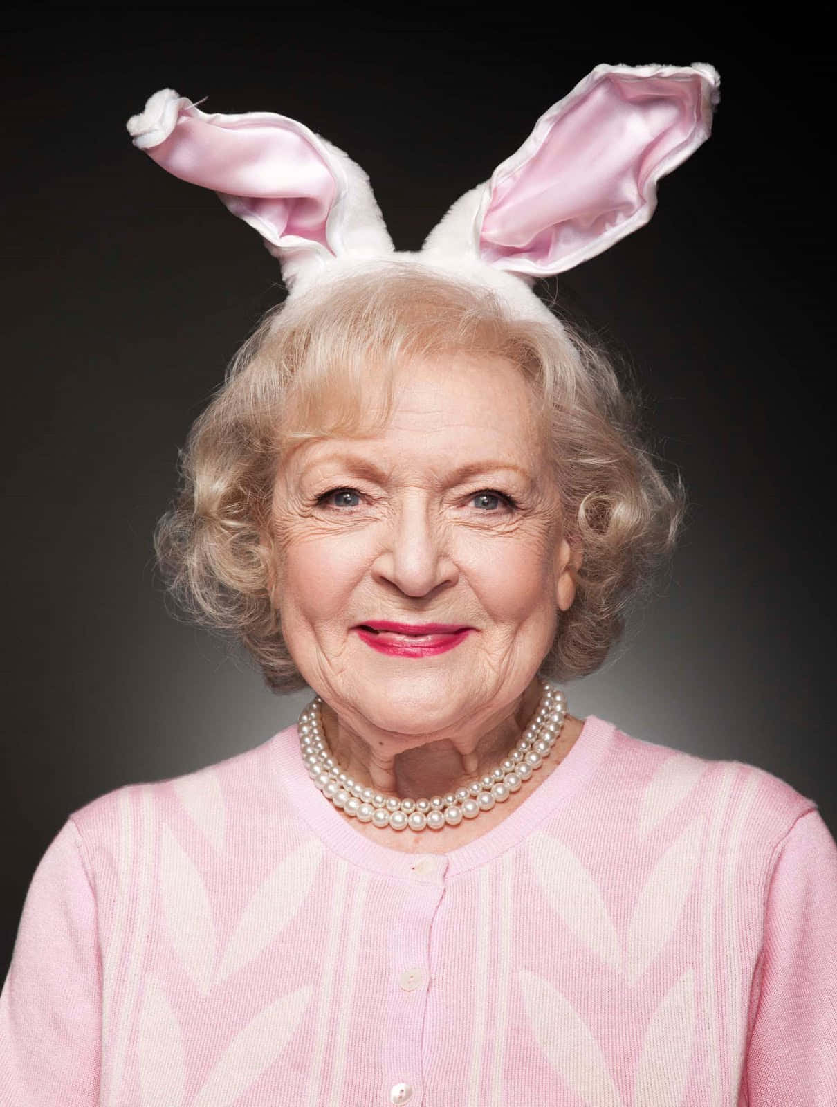 Betty White 桌布 壁紙 — 一位年長女性，金色捲髮，戴著粉紅色緞面兔耳朵和珍珠項鍊，背景為深色。