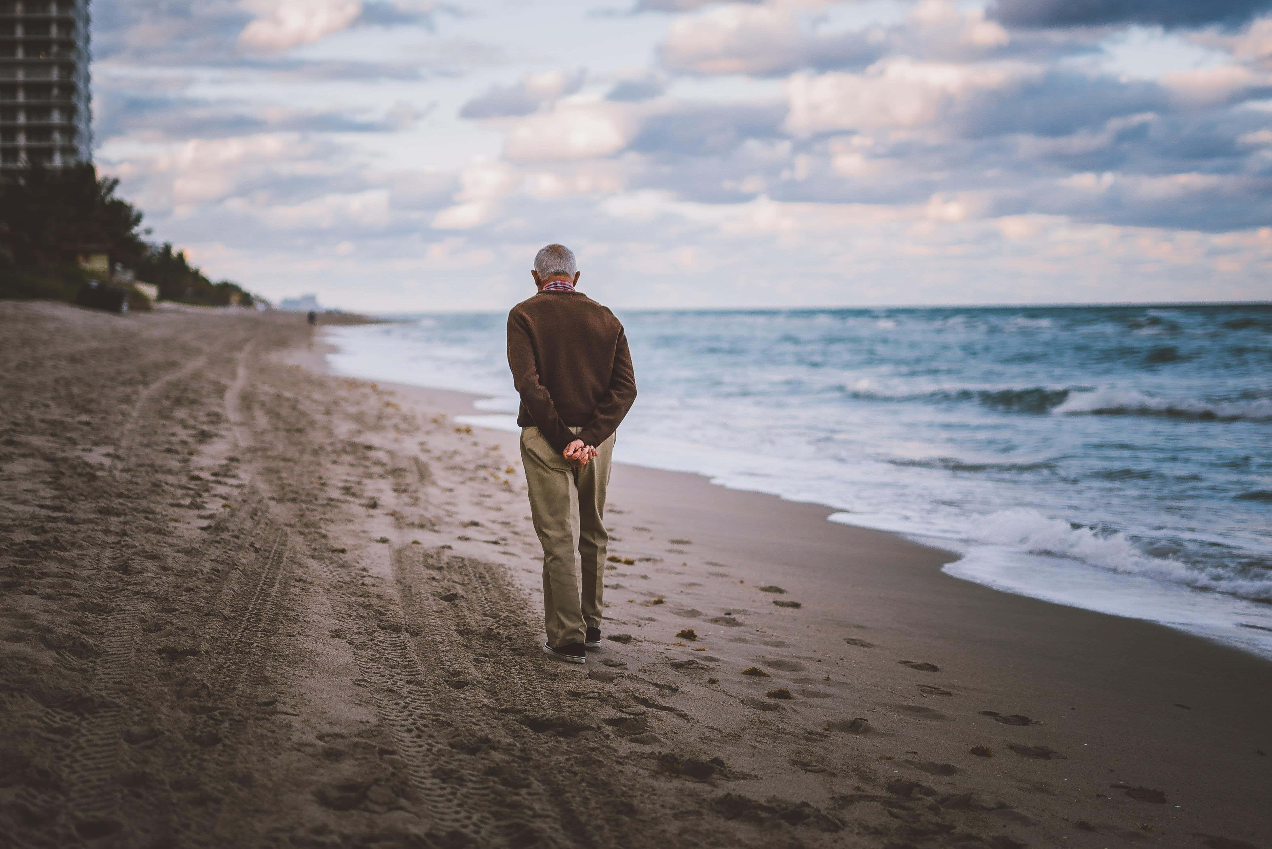 Beach Walk 桌布 壁紙 — 一位年長男子在黃昏時獨自走在沙灘上，附近有海浪拍打，頭頂上是多雲的天空。