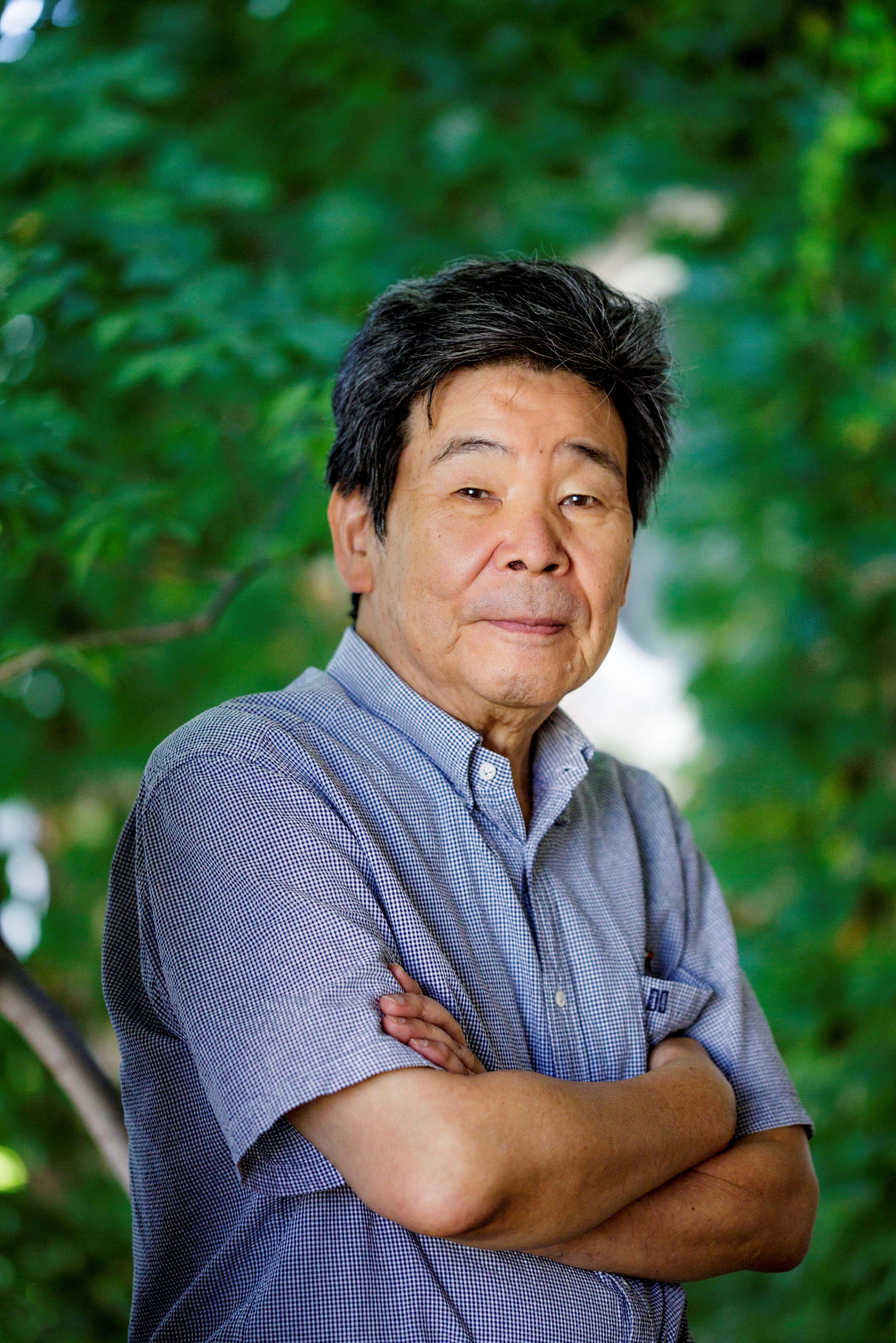 Isao Takahata 桌布 壁紙 — 一位年長男性交叉雙臂，自信地站在茂密綠色樹葉背景前。