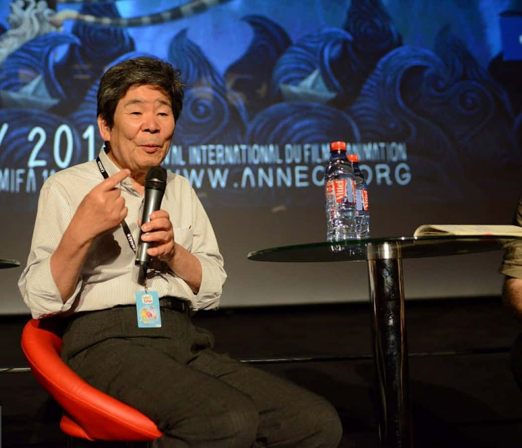 Isao Takahata 桌布 壁紙 — 一位年長男性坐在紅色椅子上，在動畫節座談會上對著咪高峰發言，背景為藍色龍。
